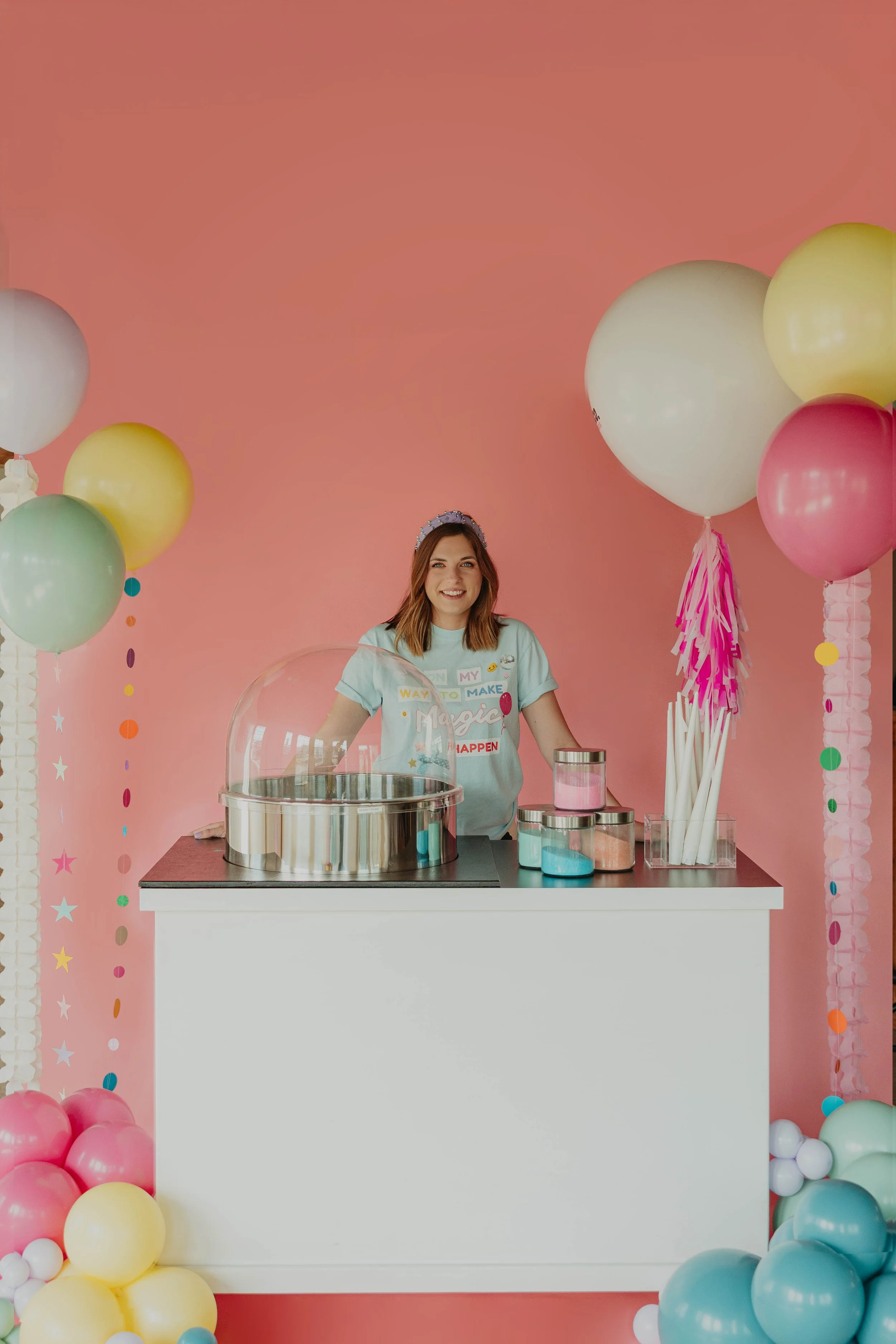 A woman stands behind a white counter decorated with pastel-colored balloons against a pink background. She has shoulder-length brown hair and is smiling, wearing a light blue shirt with colorful text and graphics. The counter has a large glass container, containers with pink, blue, and clear liquids, and a bundle of white paper straws with pink and white fringe decorations.