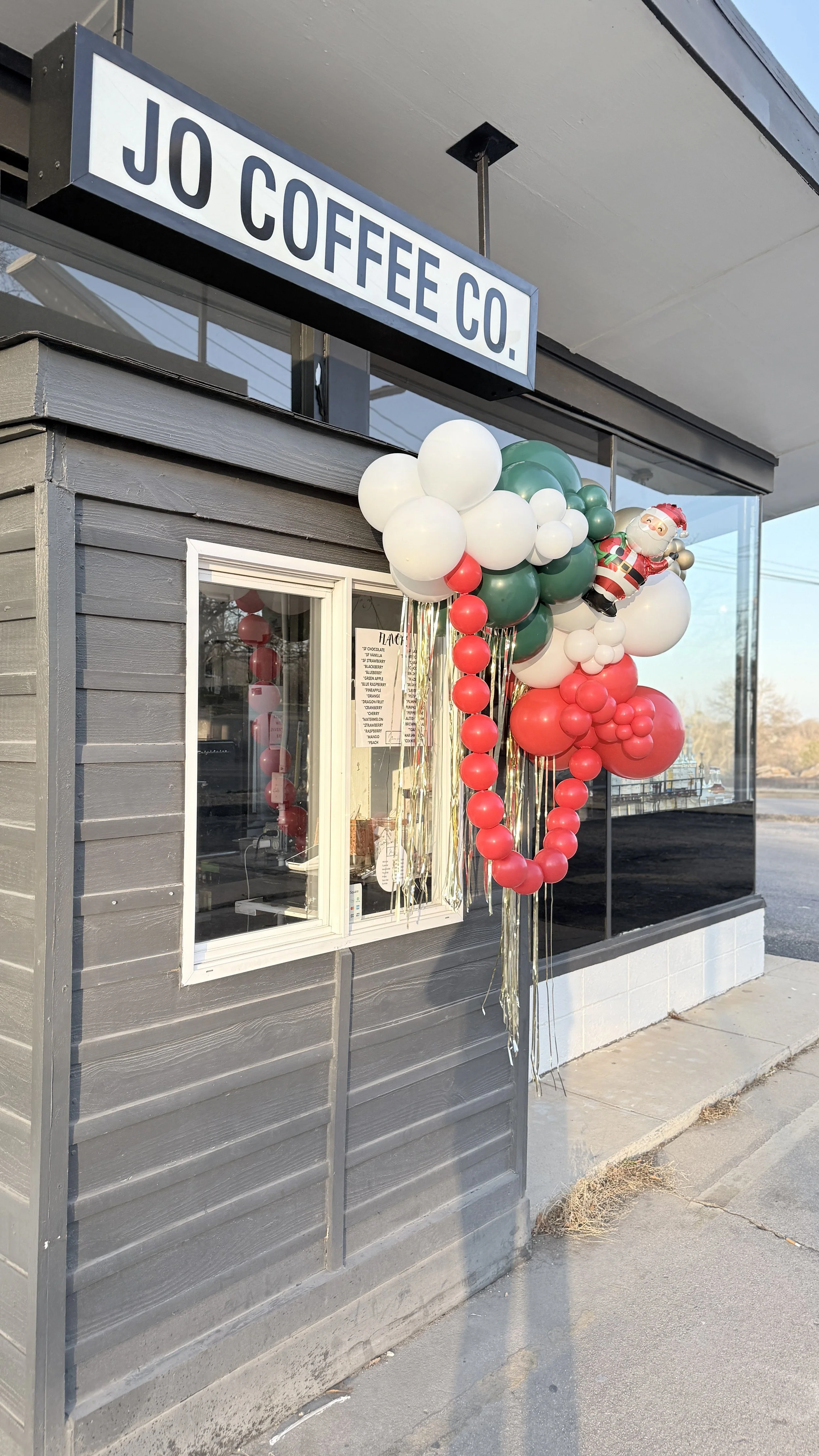 Decorations outside a small coffee shop with a balloon arrangement including white, red, and green balloons, and a Santa Claus figure, indicating a festive holiday season.