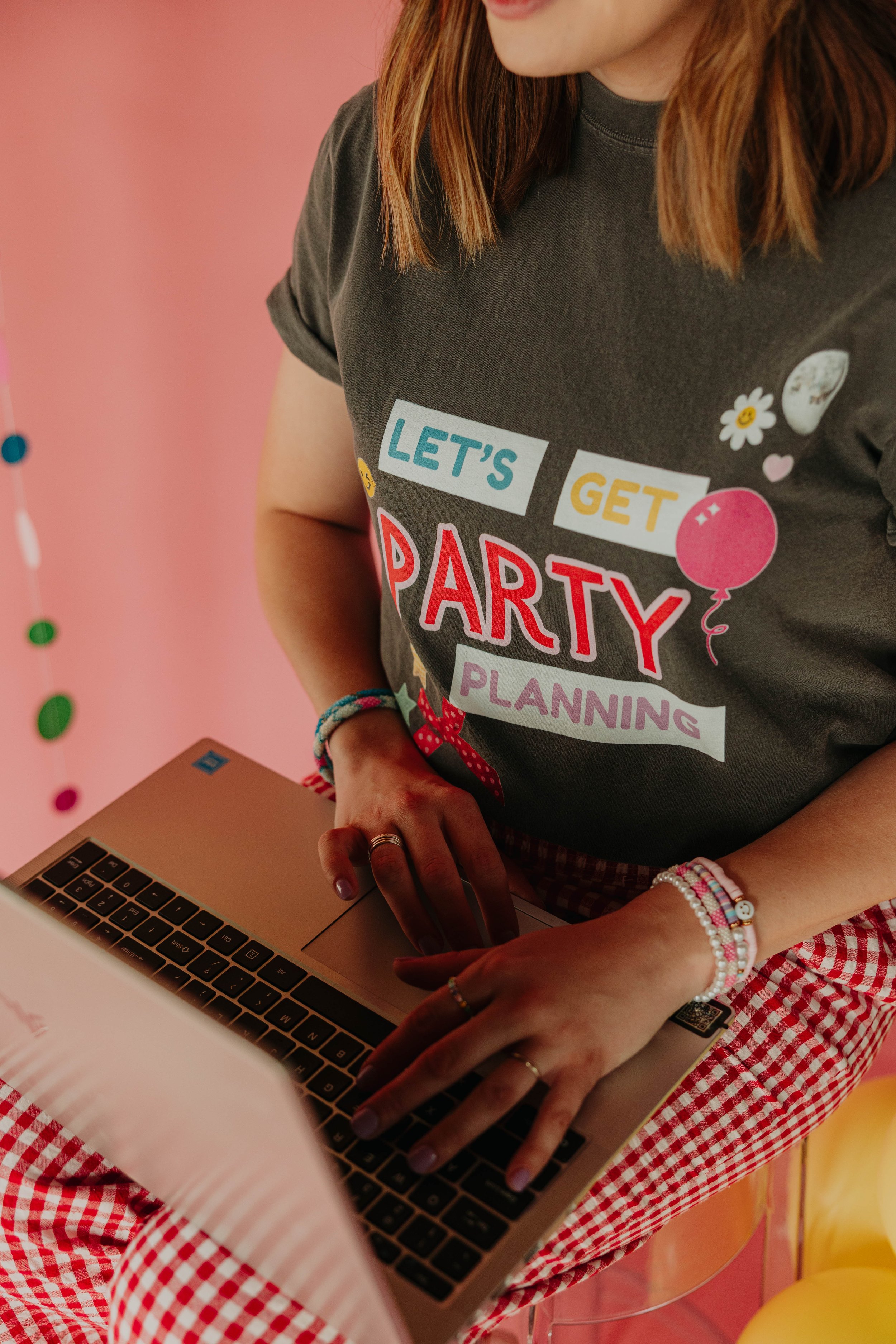Woman wearing a dark gray t-shirt with colorful text and balloons, sitting with hands on a silver laptop, in a room decorated with pink and yellow balloons.
