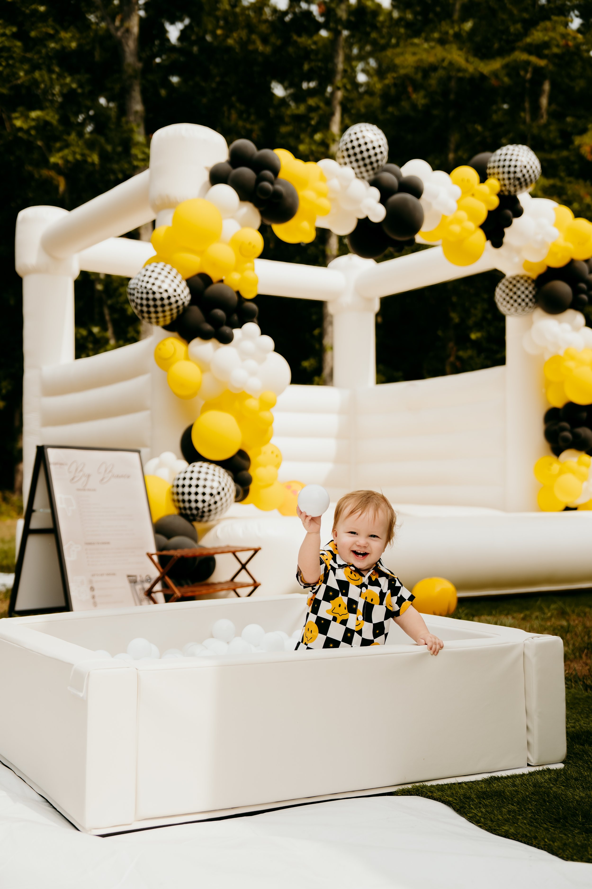 A baby with a big smile holding a white ball, standing in a white ball pit at an outdoor party with black, yellow, and white balloon decorations and a white inflatable structure in the background.