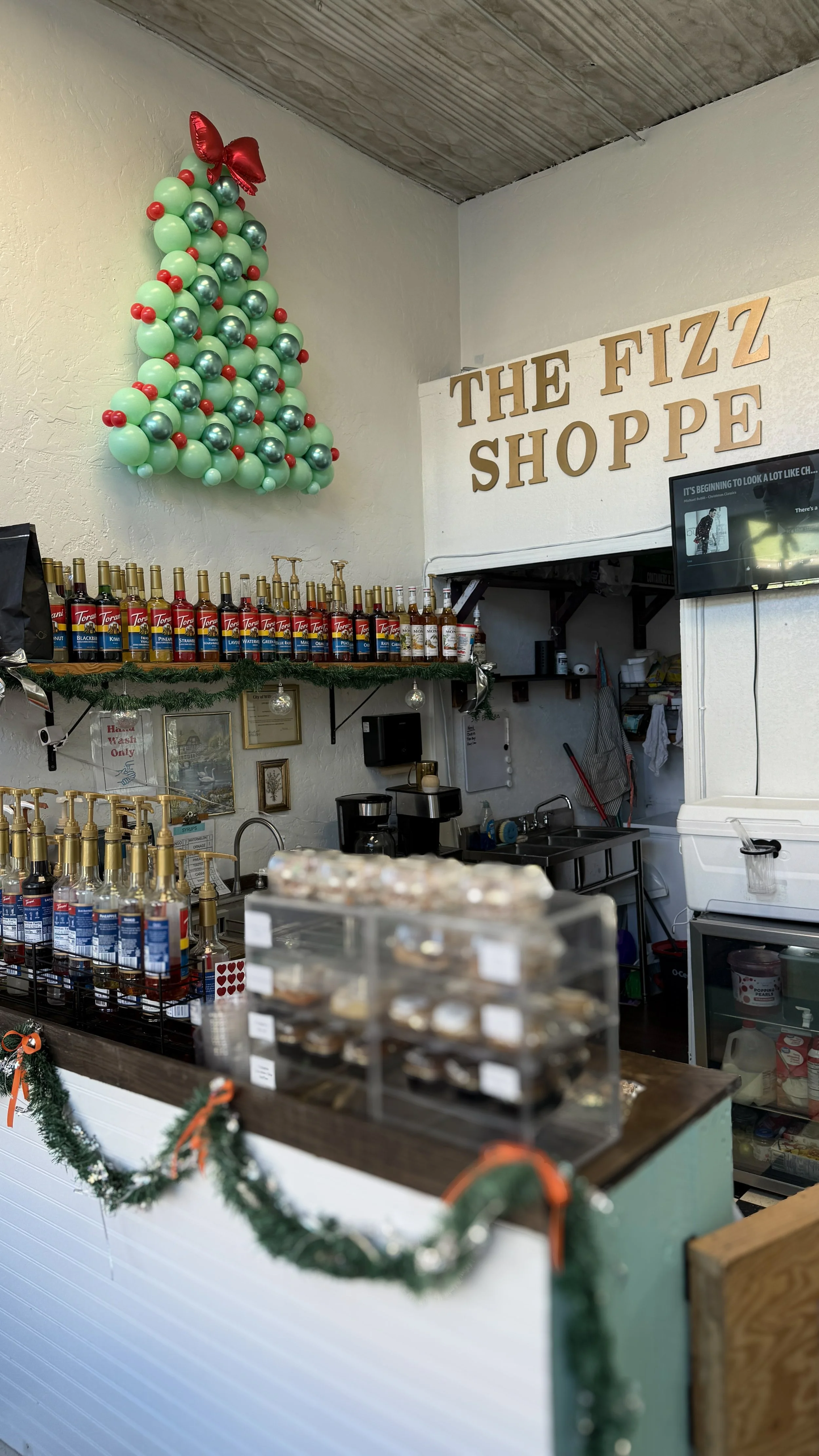 Interior of a shop decorated with Christmas ornaments, including balloons arranged in the shape of a Christmas tree with a red balloon bow on top. The shop has bottles of flavored syrups on a shelf, framed pictures, a sign reading 'The Fizz Shoppe,' 