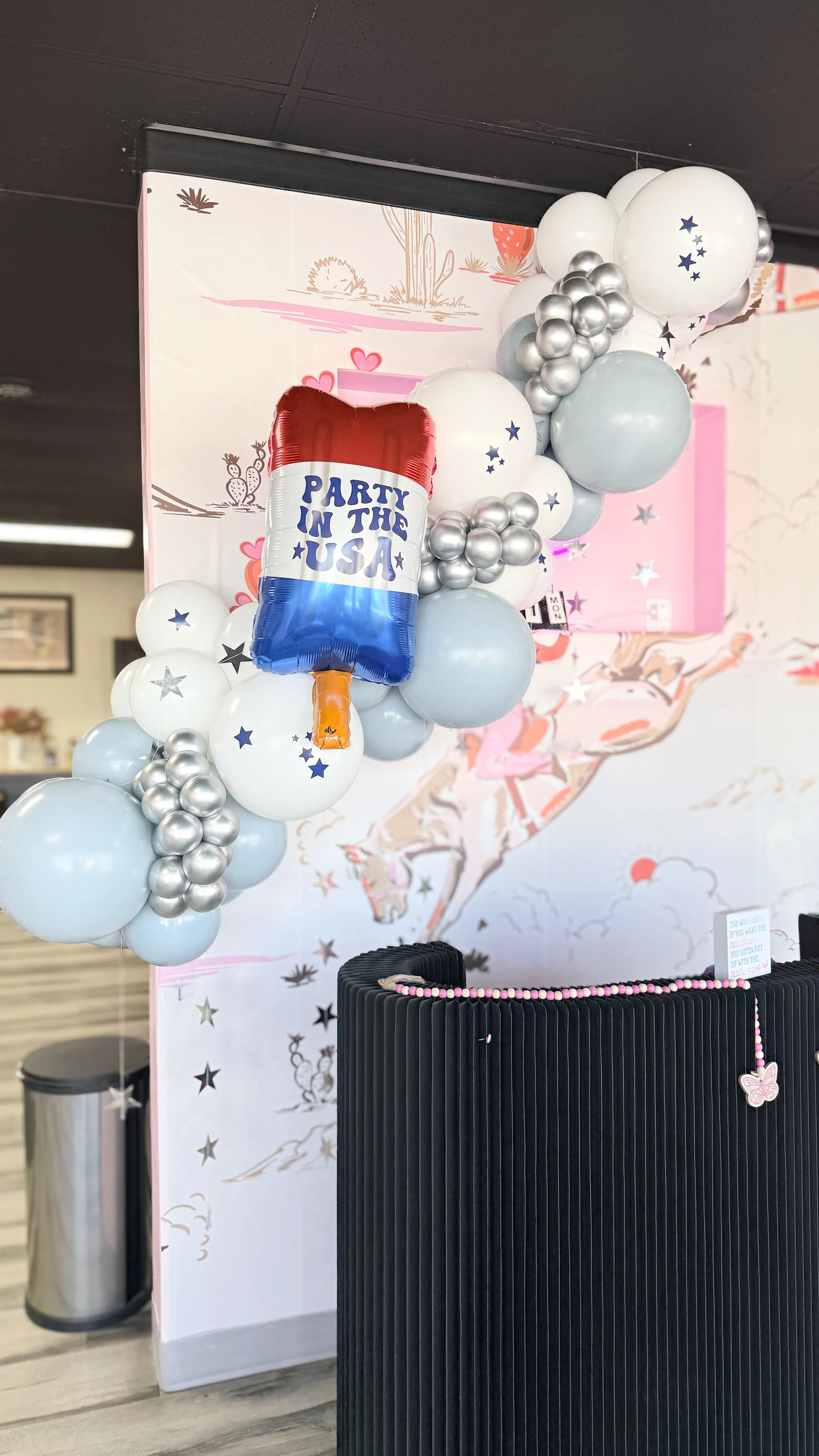 Balloons with a USA flag design and a message reading 'Party in the USA' hanging among other white, silver, and light blue balloons, with a backdrop featuring desert landscape and a pink fox illustration.