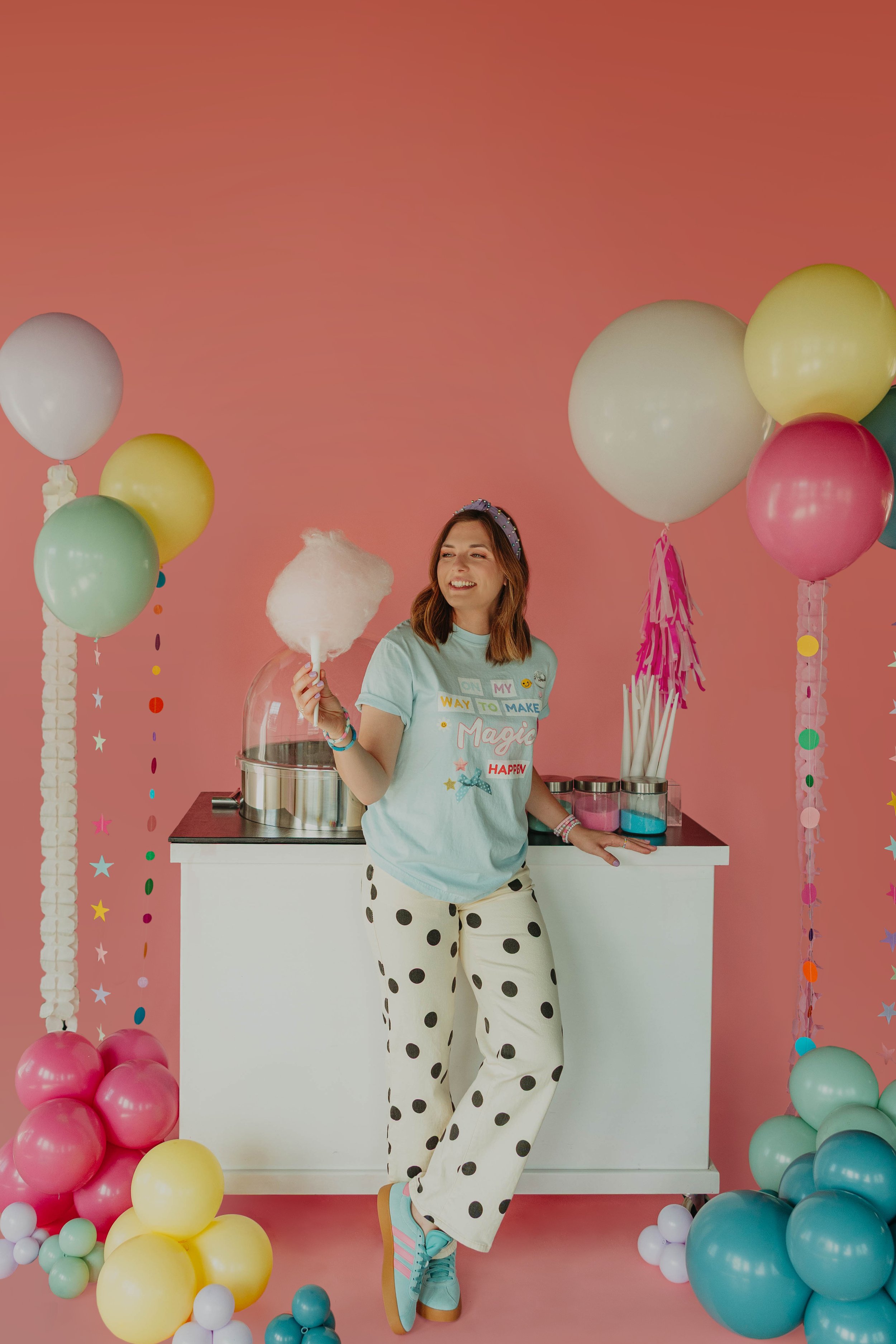 A woman standing in a decorated party setting with balloons and colorful streamers, holding pink cotton candy, smiling and wearing polka dot pants, a graphic T-shirt, and colorful sneakers.