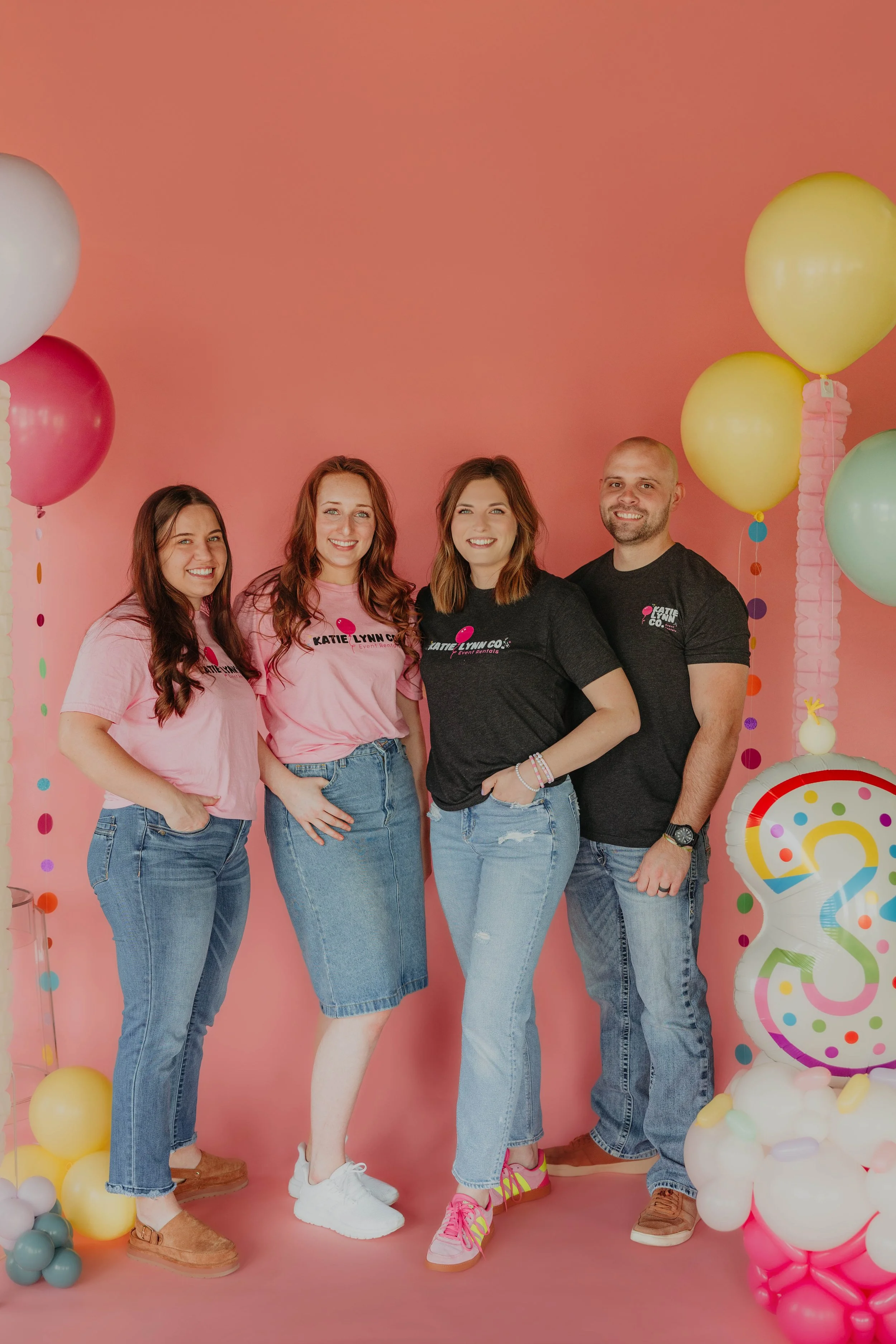 Five smiling people standing against a pink background with colorful balloons, wearing casual clothing including pink and black t-shirts with a logo and jeans.