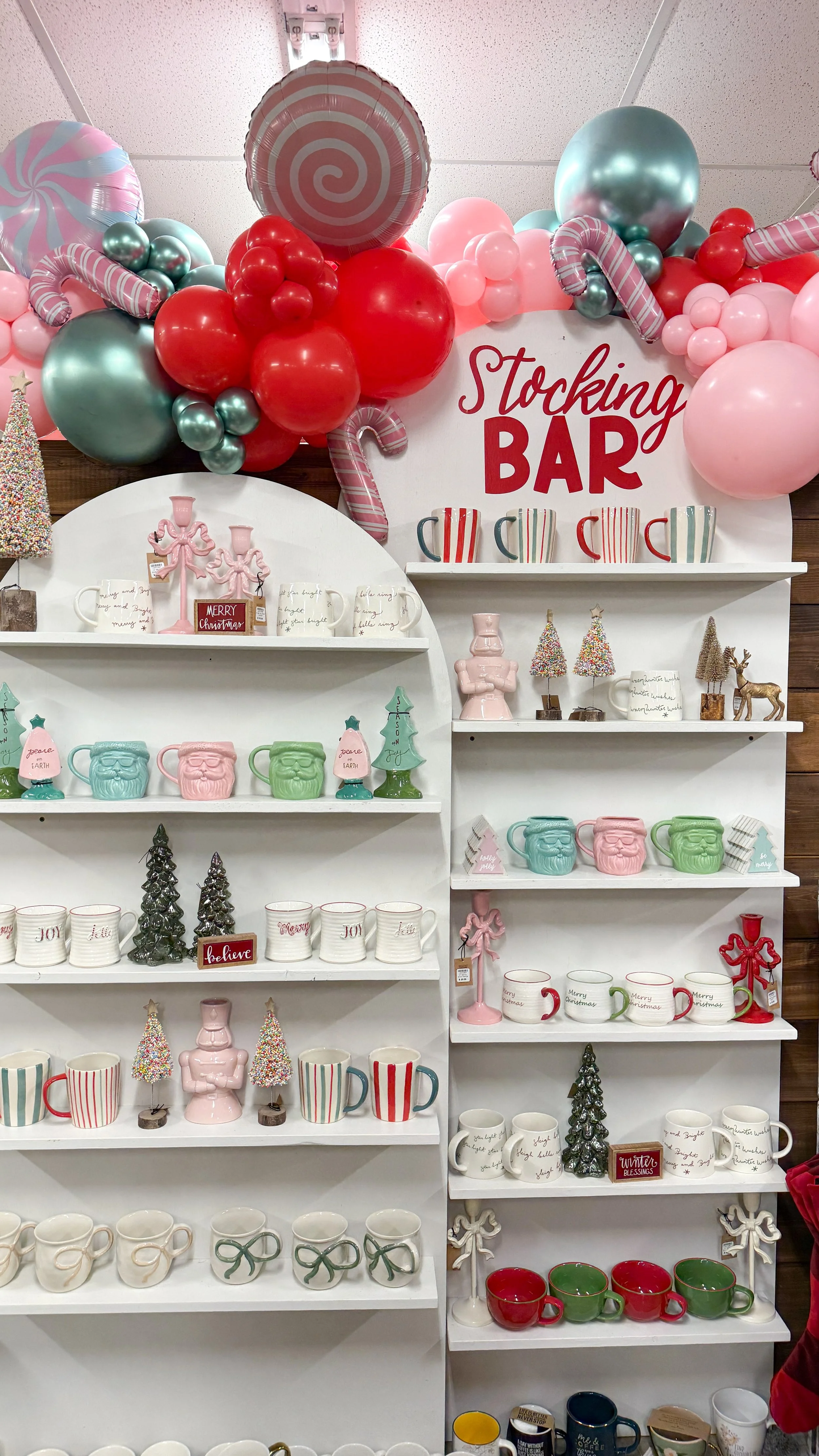 Decorative store display with holiday-themed ceramics and ornaments, including Christmas trees, reindeer, and mugs with festive messages, set against a backdrop of pink, red, green, and white balloons.