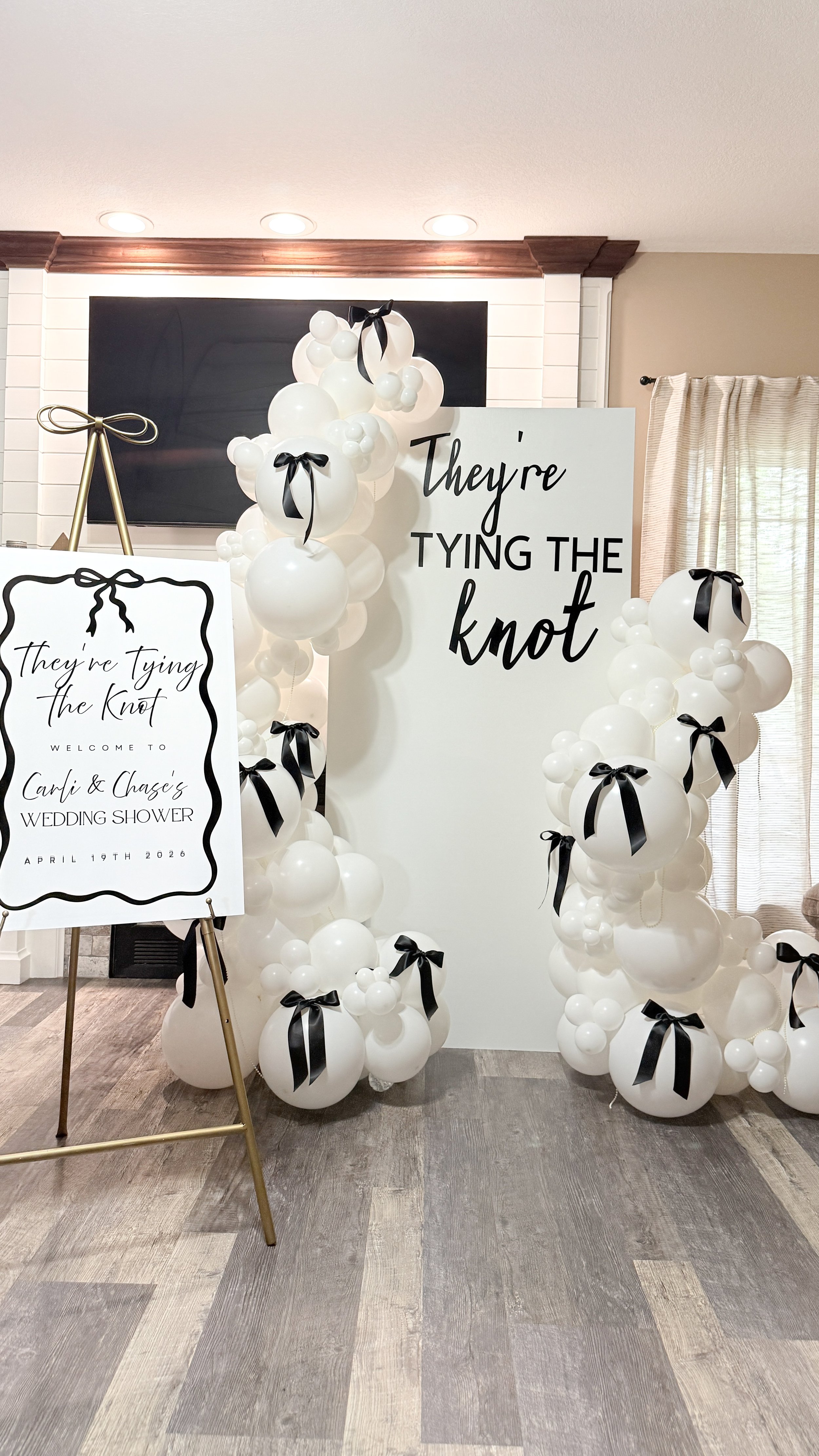 Decorative wedding shower setup with white balloons tied with black ribbons, a large sign with the phrase "They're Tying the Knot," and a welcome sign for Cari and Chase's wedding shower on April 19th, 2026.