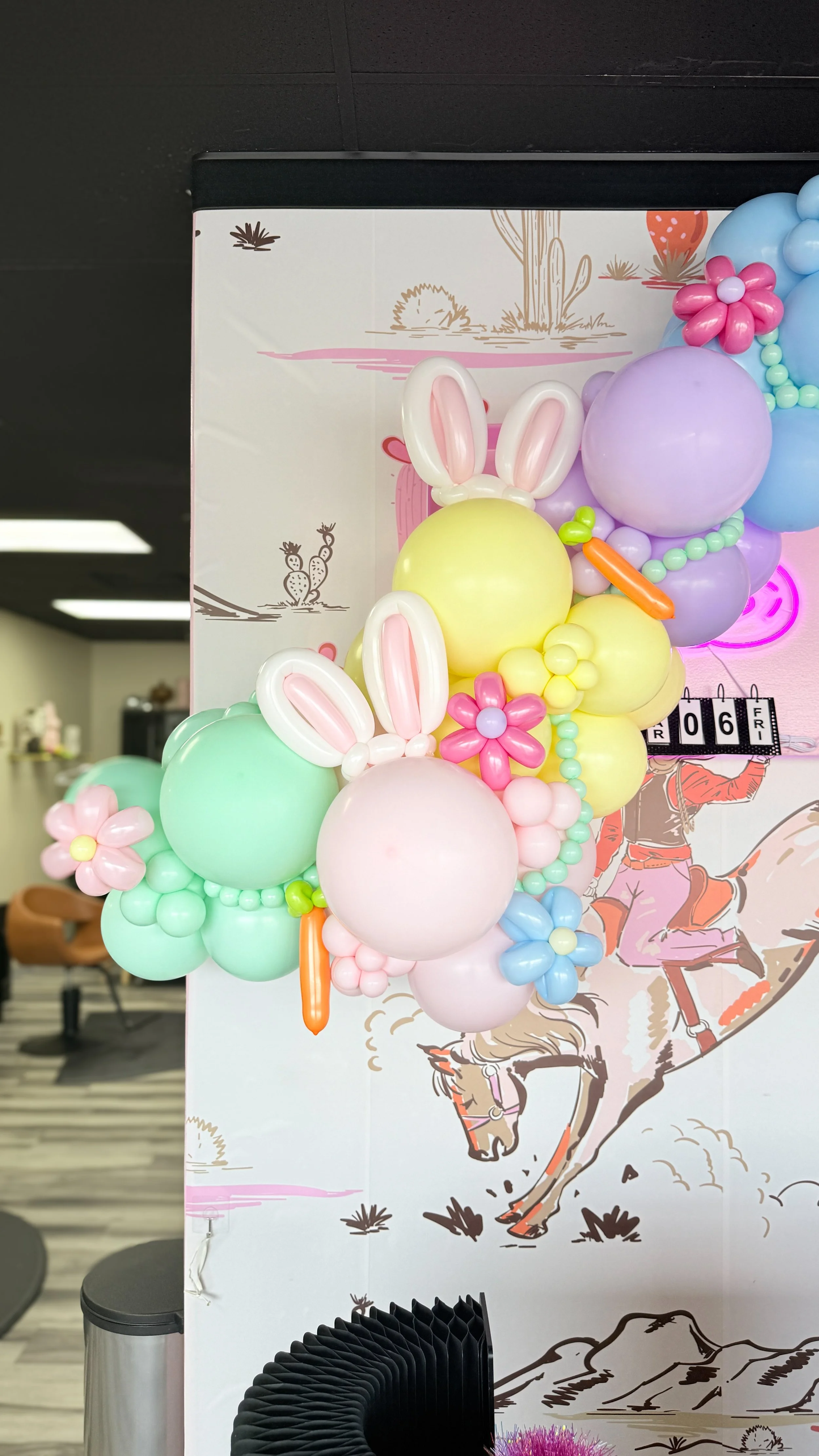 Decorative balloon arrangement featuring pastel-colored balloons shaped like bunny ears, flowers, and carrots, attached to a backdrop with a desert scene illustration.