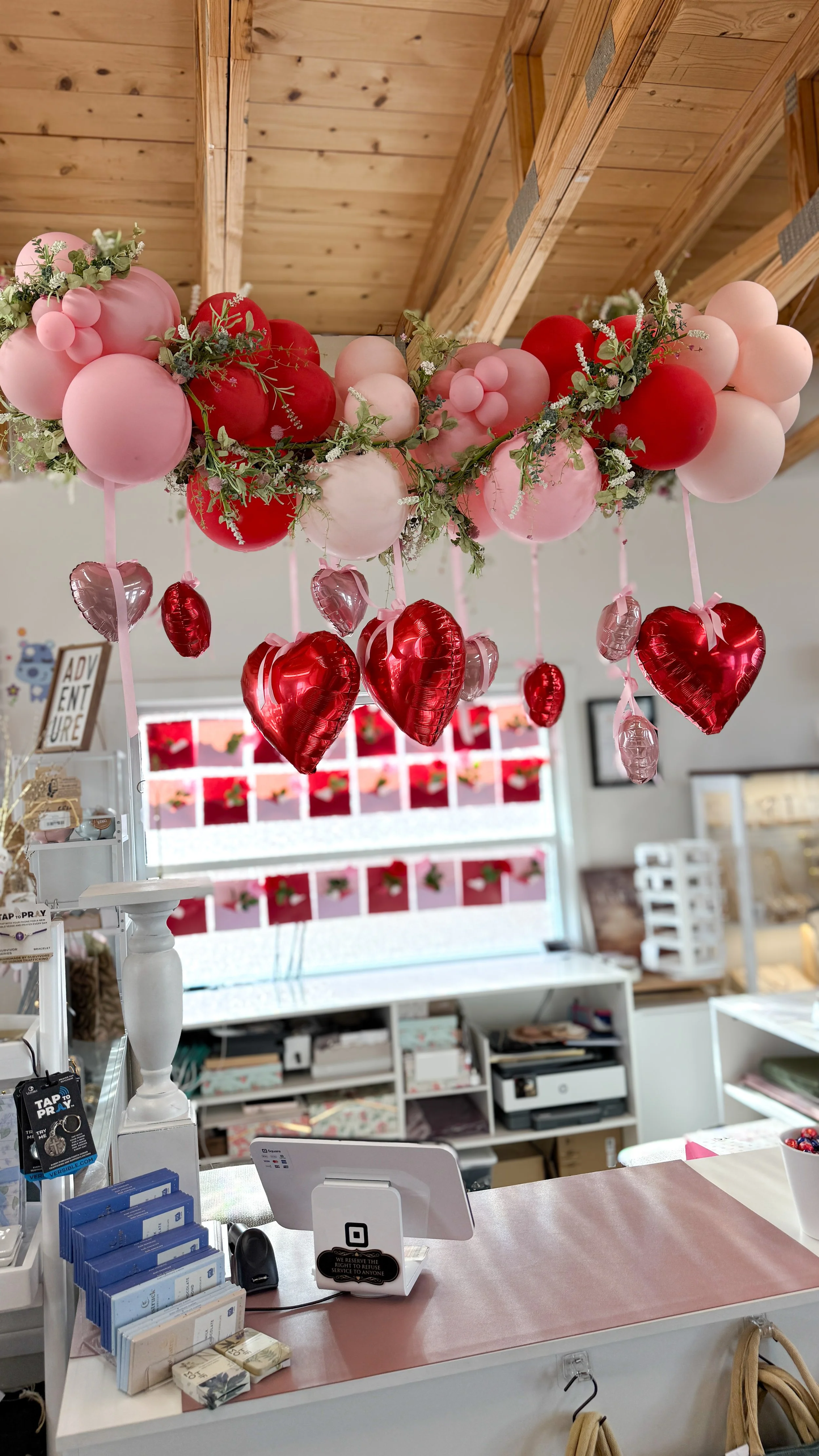 Decorative balloon arrangement with pink and red balloons and greenery above a store counter.