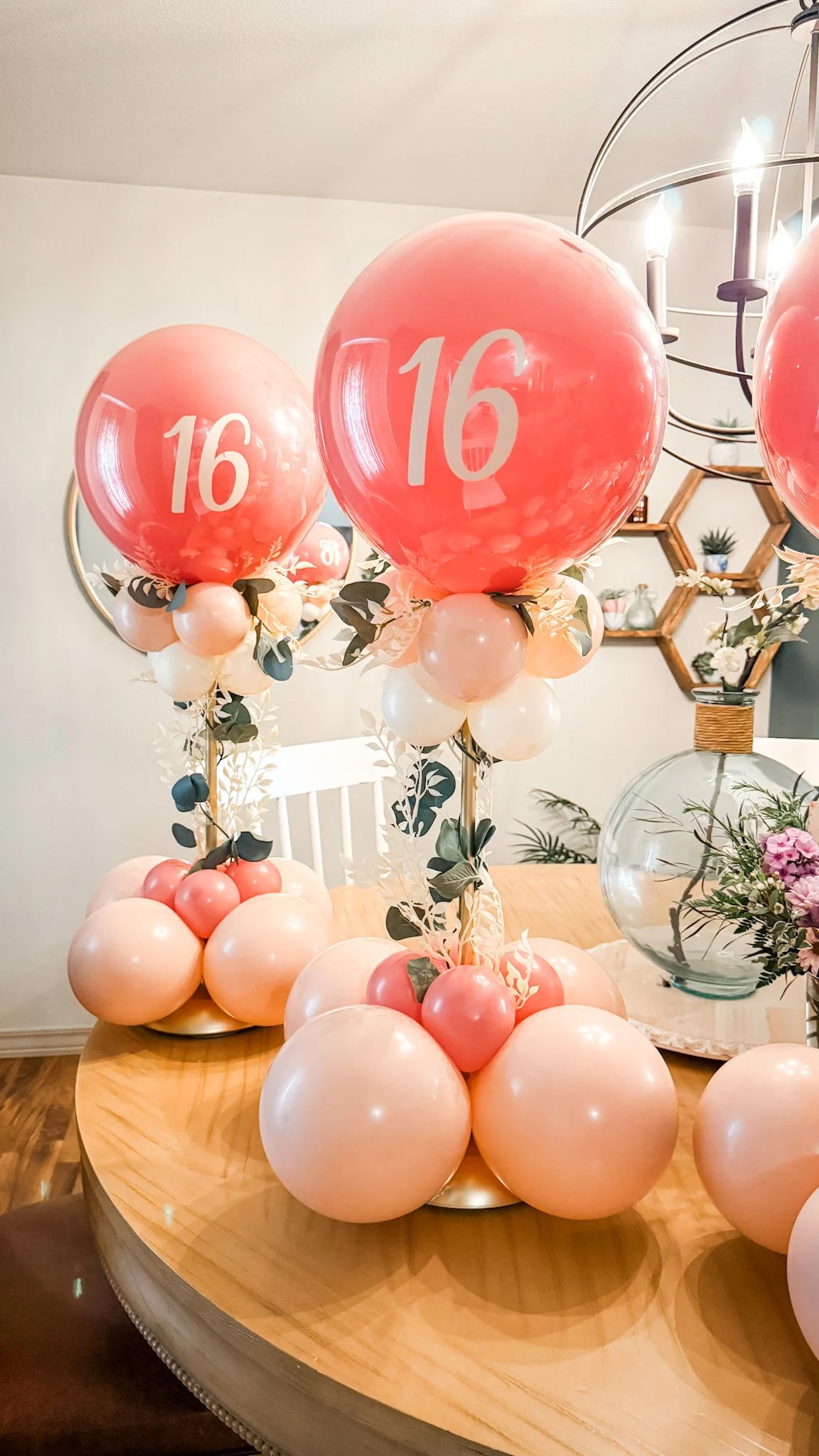 Decorative balloon arrangements with pink and white balloons, featuring large pink balloons with white number 16, on a wooden table for a 16th birthday celebration.
