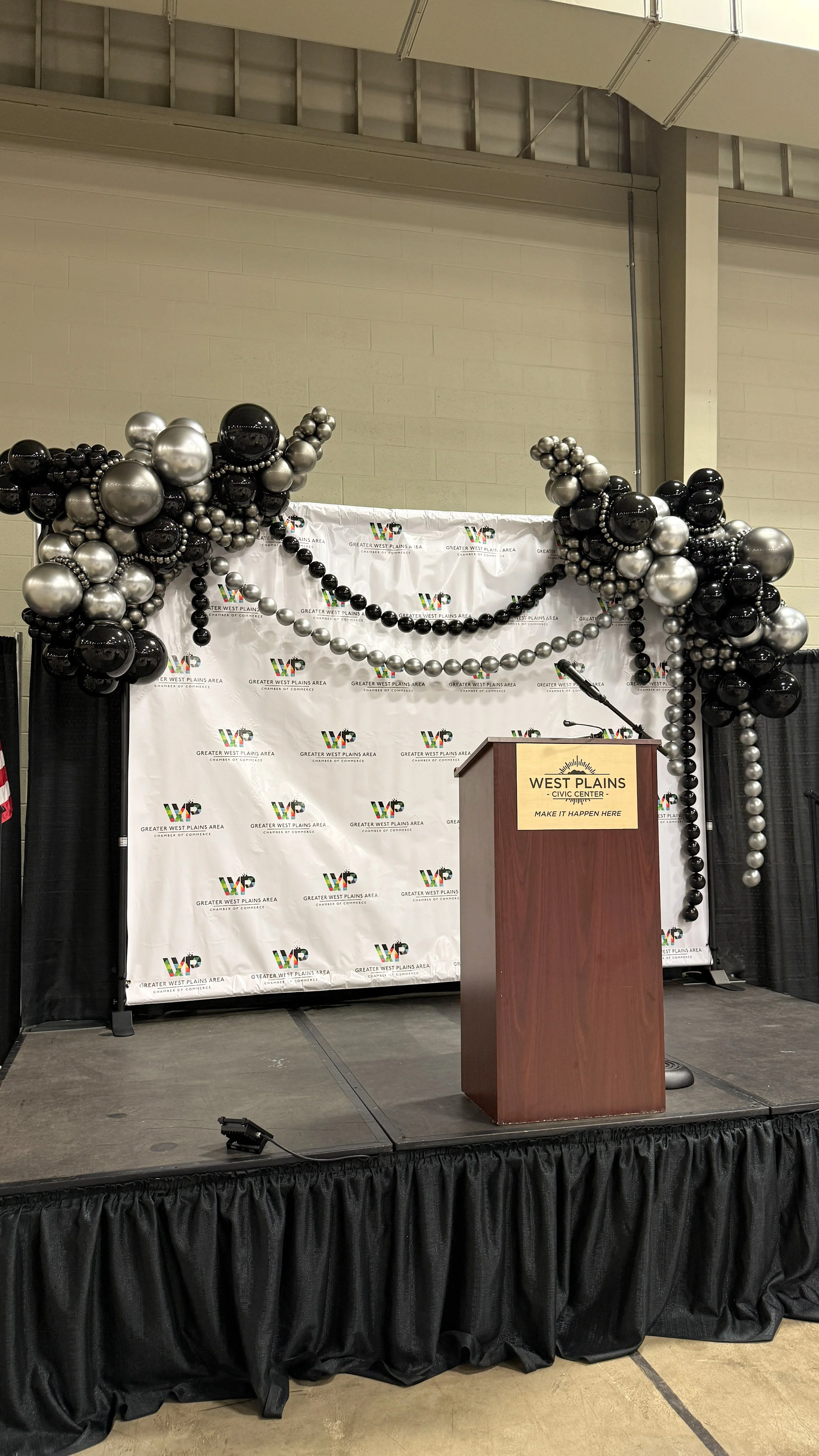 A stage decorated with black, silver, and pearl-colored balloons in front of a backdrop with logos of the Greater West Plains Area Chamber of Commerce. There is a wooden podium with a sign that reads 'West Plains Civic Center, Make It Happen Here' and a microphone on top.