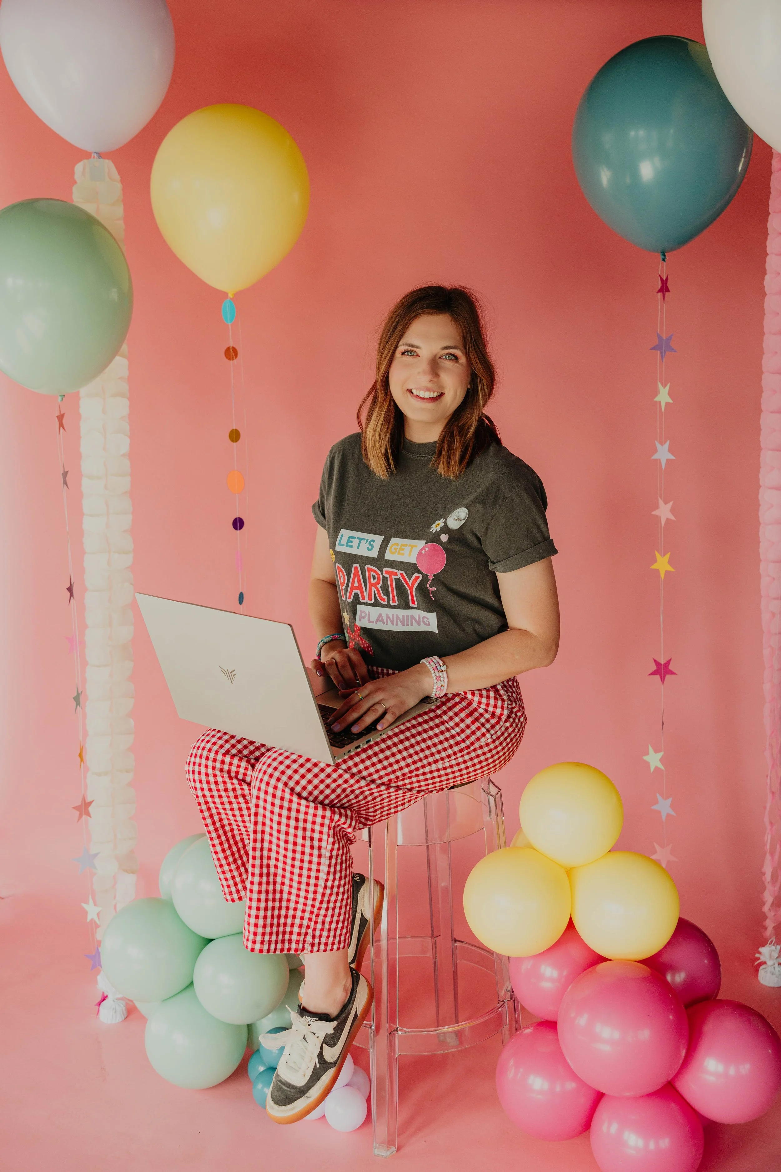A young woman with brown hair sitting on a clear stool surrounded by pastel-colored balloons in pink, yellow, green, blue, and white, against a pink background decorated with star-shaped garlands. She is smiling, wearing a black T-shirt with a birthday-themed print and red-and-white checkered pants, holding her laptop on her lap.