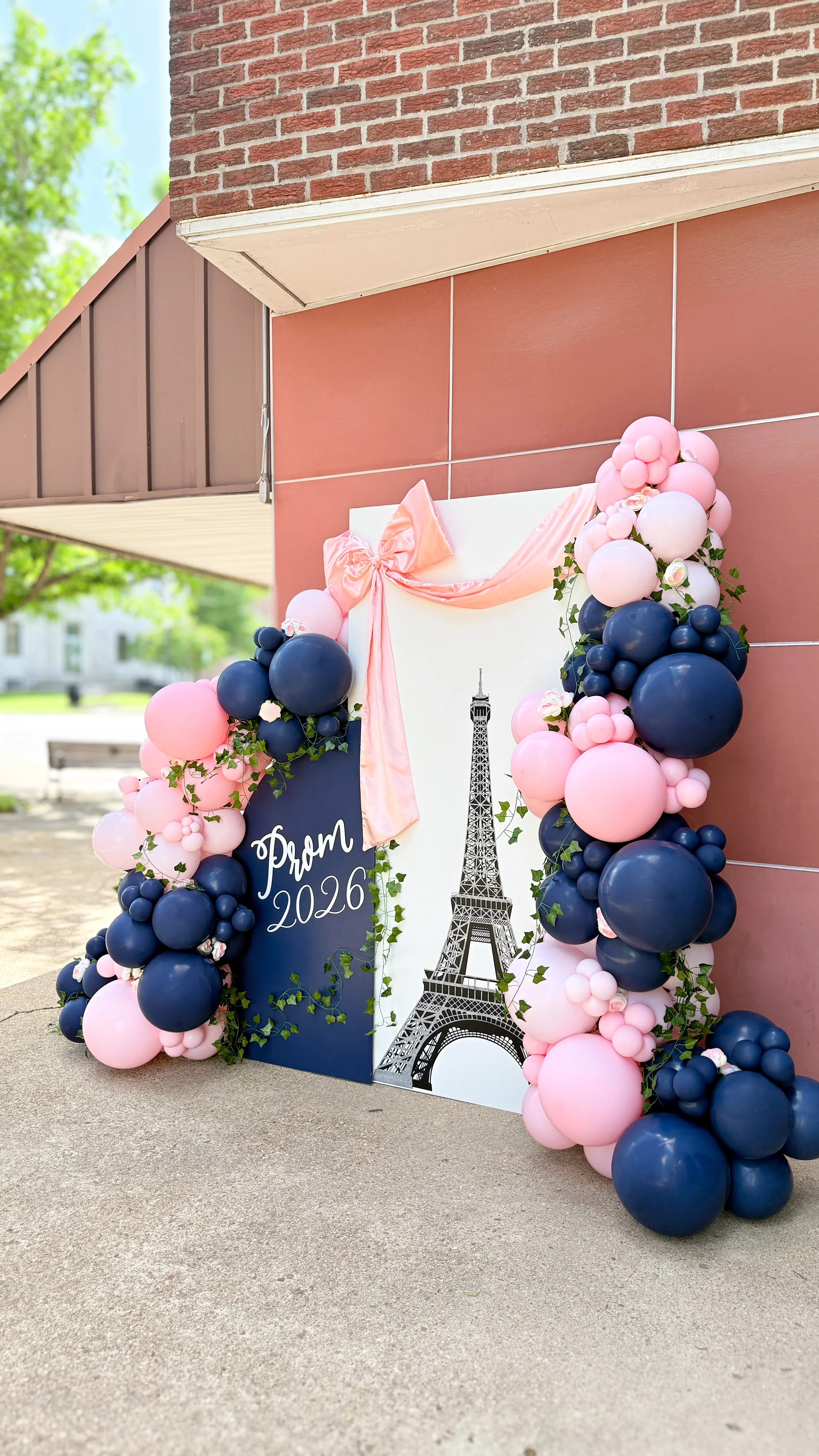 Pink and navy blue balloon garland decorated backdrop with greenery, outdoors, against a red brick wall and concrete floor 
