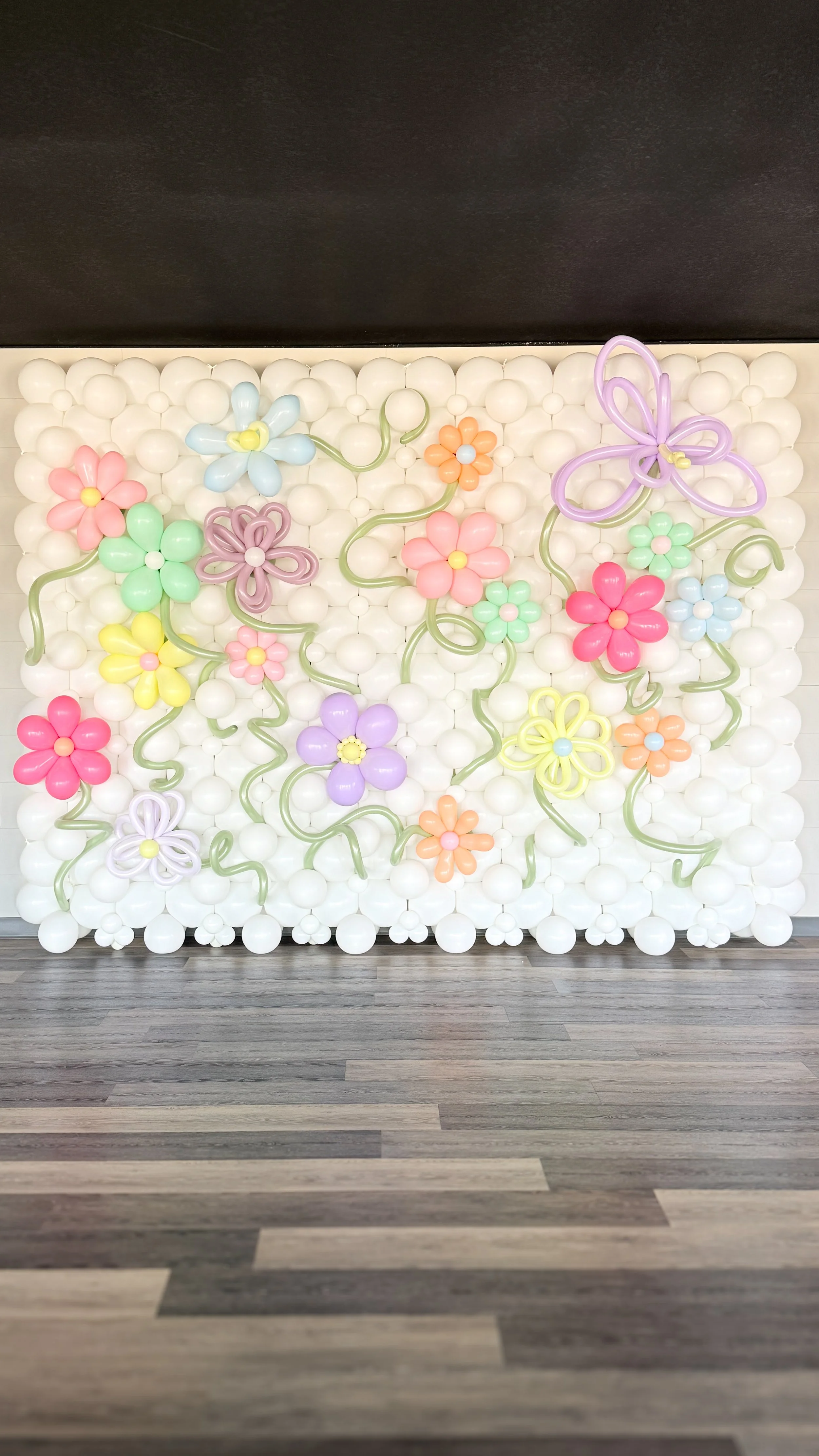 Colorful balloon art wall decorated with various balloon flowers and a large butterfly, set against a background of white balloons, with a wooden floor in front.