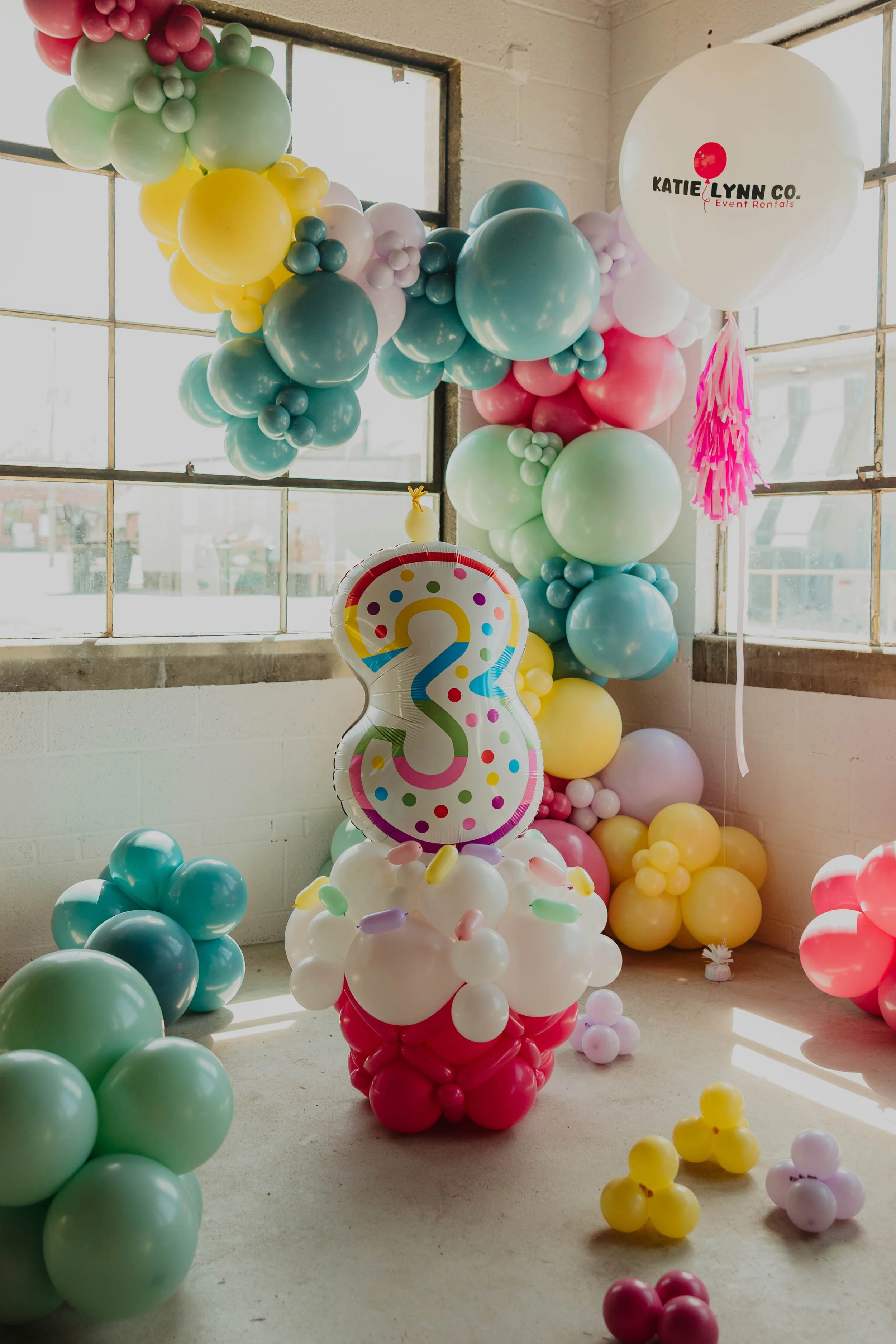 A woman wearing a white T-shirt that says 'In my balloon babe era" with pink and purple balloons around her.