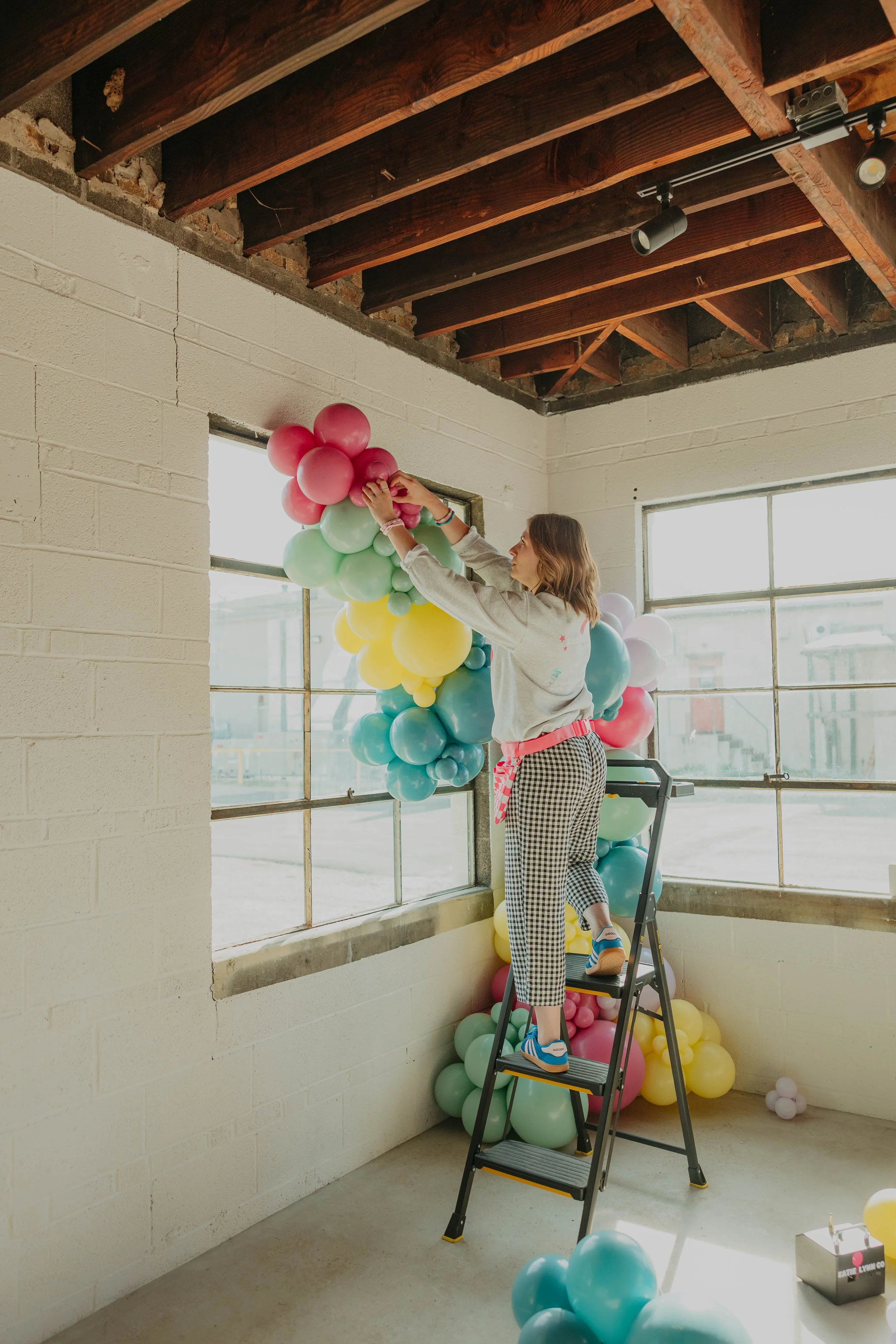 A woman with shoulder-length light brown hair, smiling, sitting on the floor with pastel-colored balloons around her, holding two yellow balloons in front of her, with a balloon pump and a cluster of balloons behind her.
