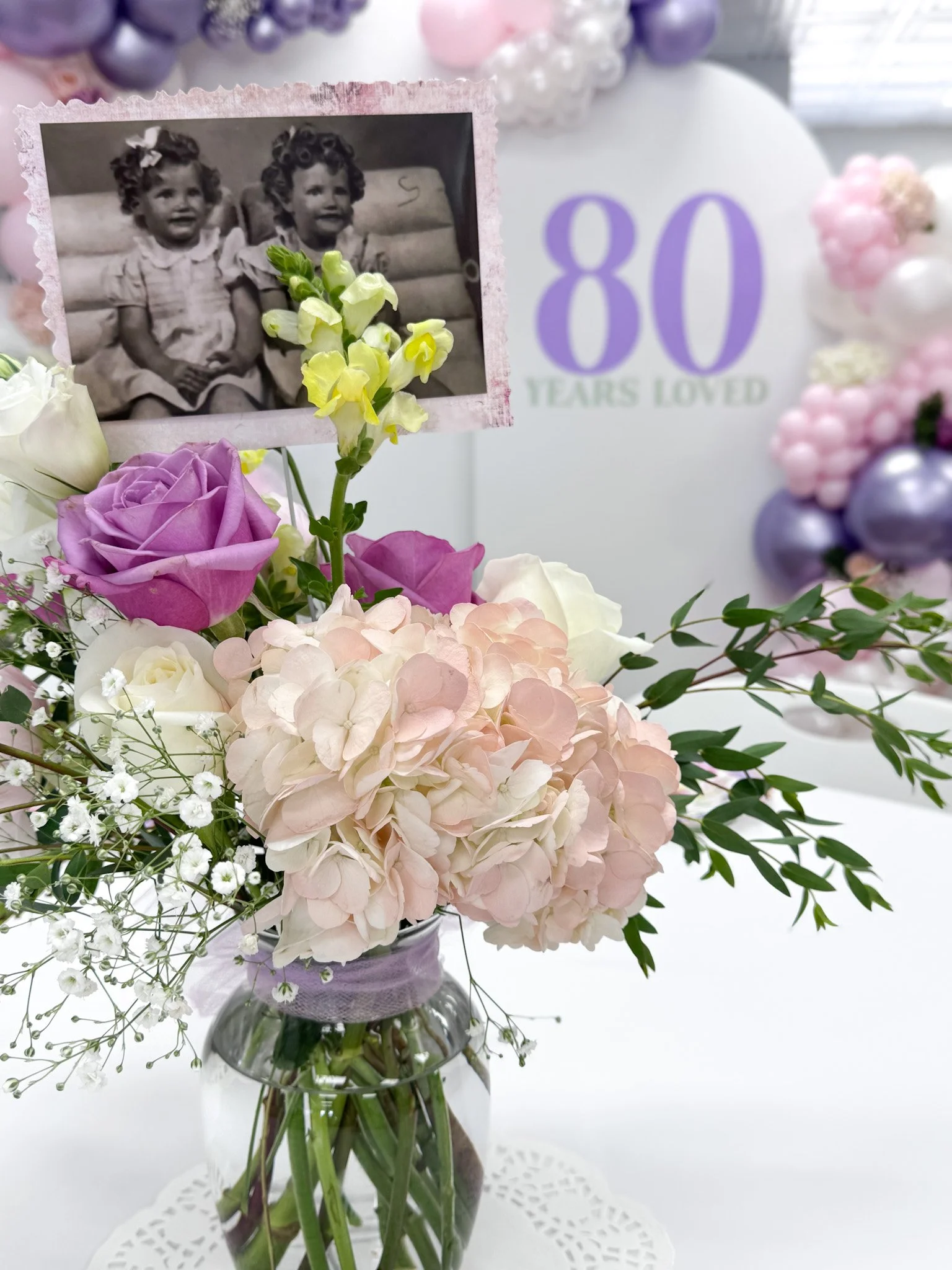A floral arrangement with pink roses, white roses, lavender hydrangeas, small white filler flowers, and greenery in a glass vase. A black and white photograph of two young girls is attached to a stick in the arrangement. In the background, a sign says '80 Years Loved' with balloons around it, indicating a celebration or milestone.
