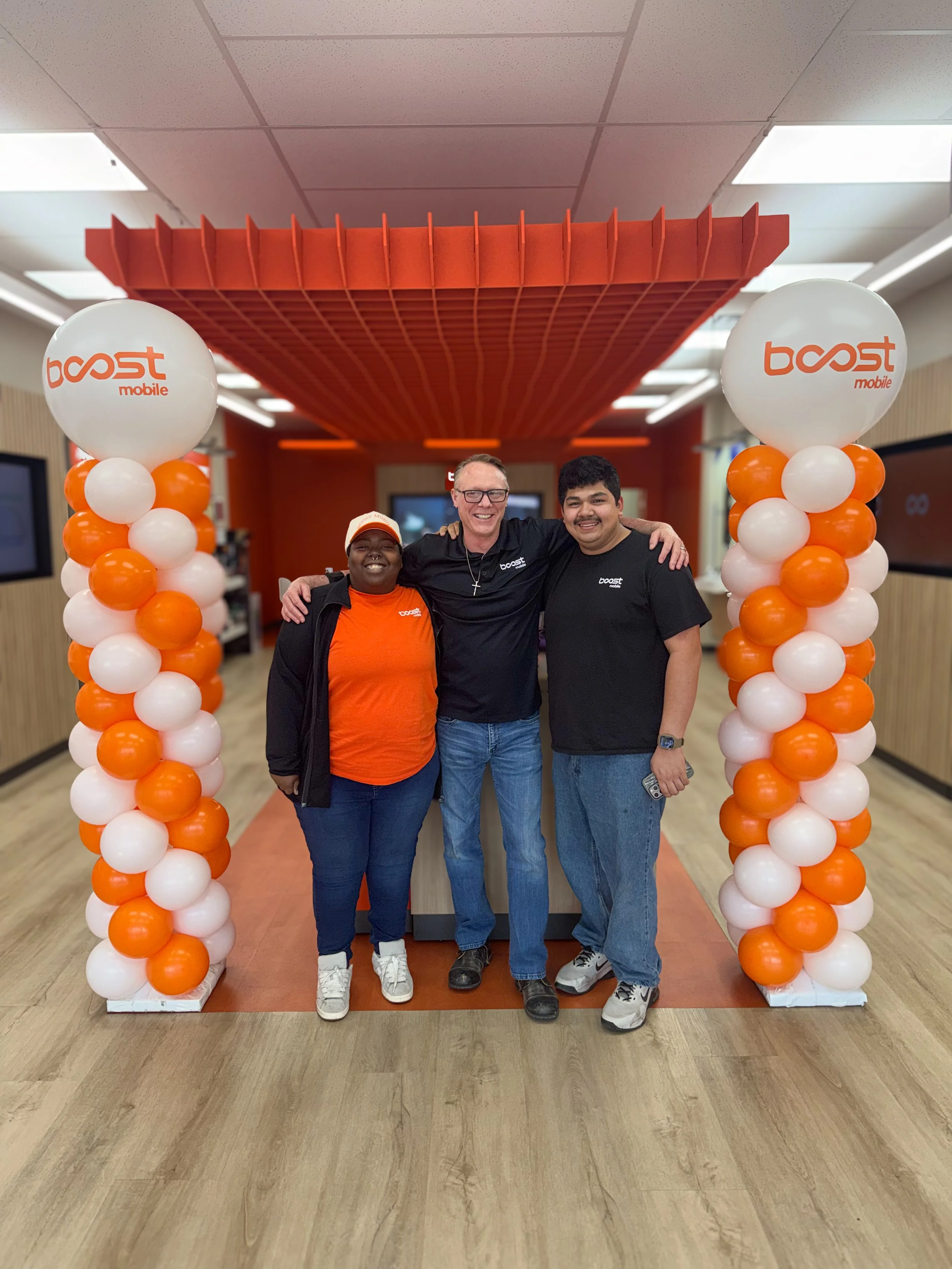 Three people standing under a balloon arch with orange and white balloons. Two women and one man smiling, with the woman on the left wearing an orange shirt and a cap, the man in the middle wearing glasses and a black shirt, and the woman on the right wearing a black shirt. They are indoors, at a Boost Mobile event or celebration.