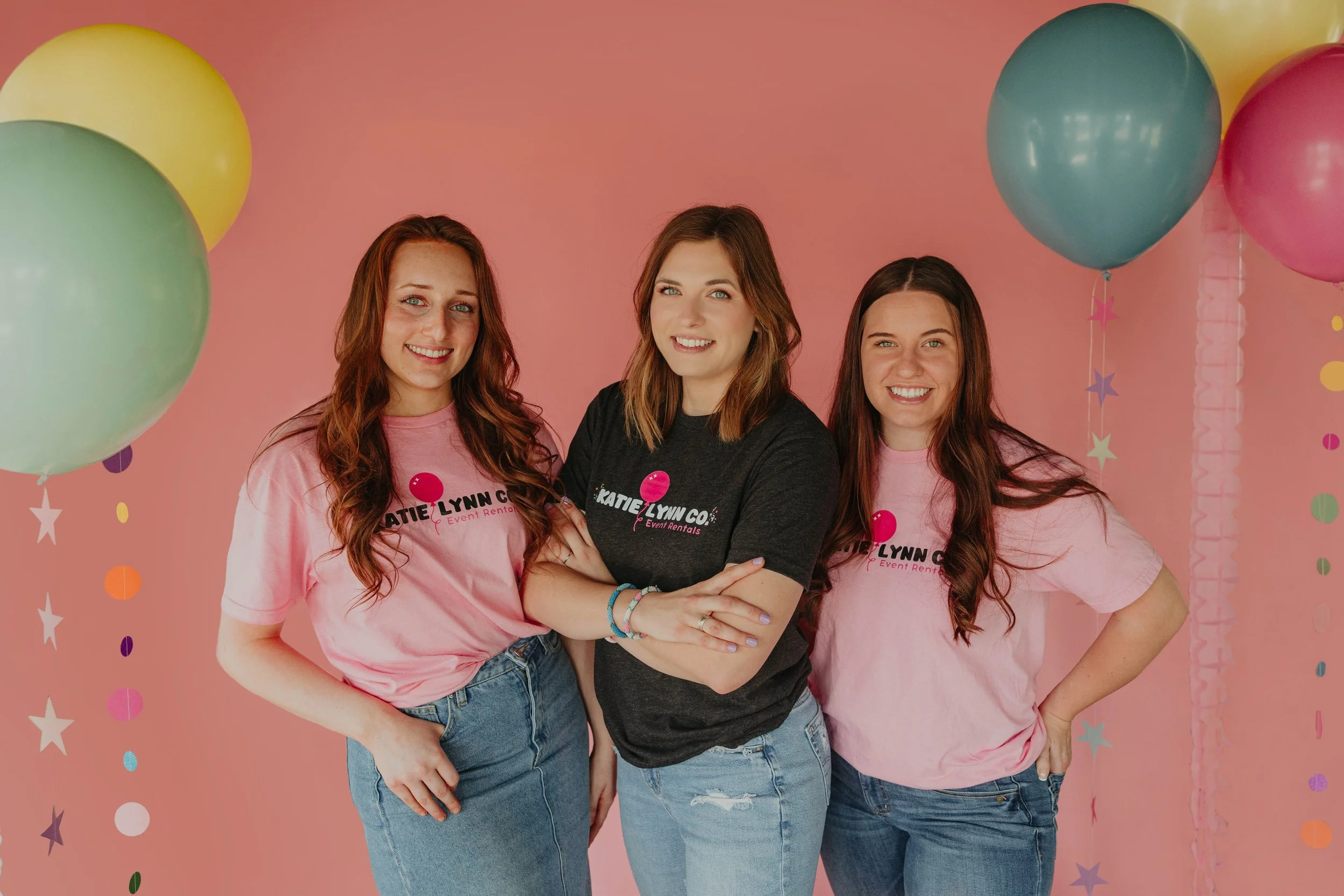 Three women standing close together smiling against a pink background with colorful balloons and confetti.