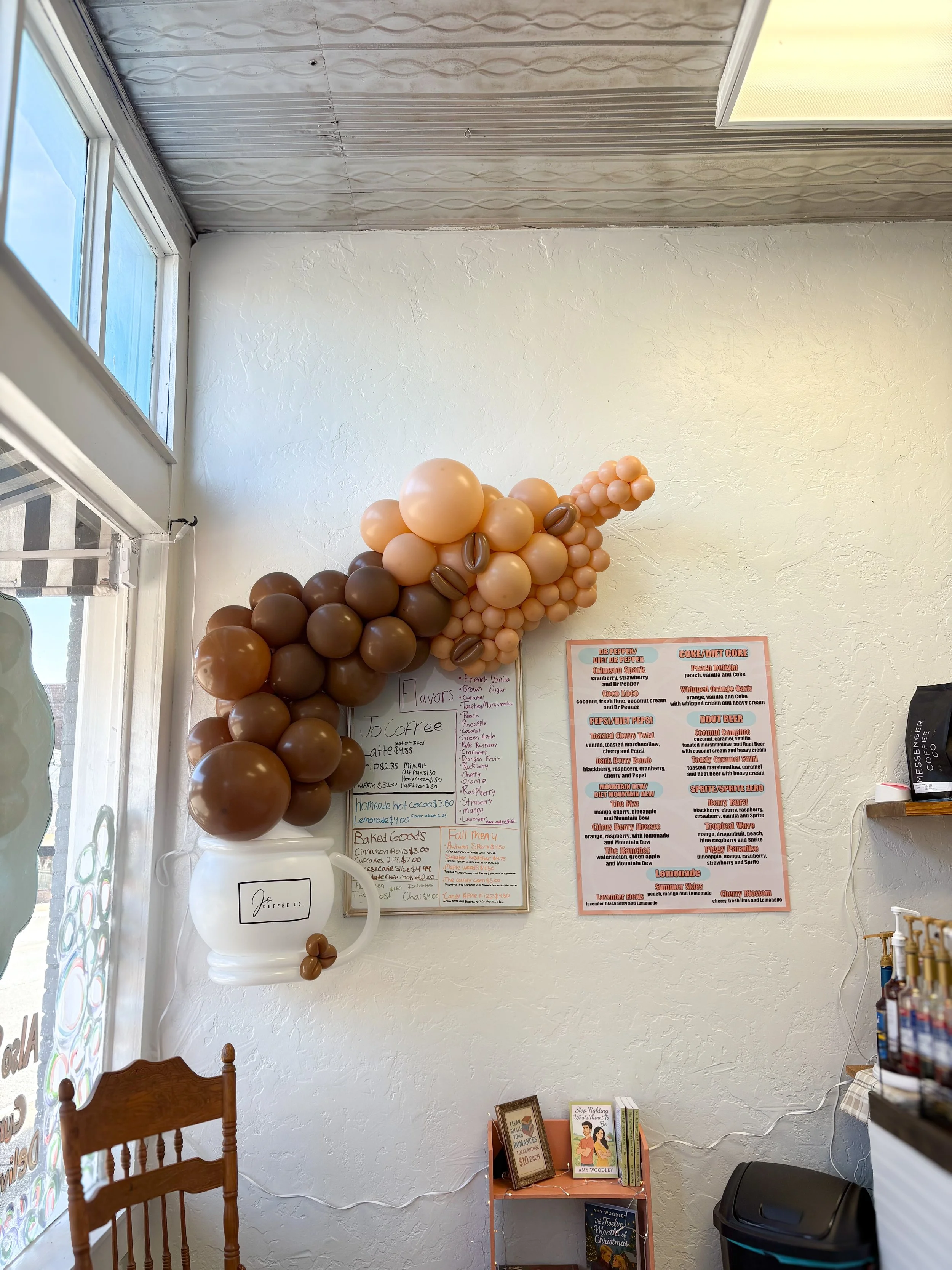 Interior of a bakery or coffee shop with a balloon decoration, a menu board on the wall, a small orange table with books, and a chair. The ceiling is metallic, and there's a window letting in natural light.