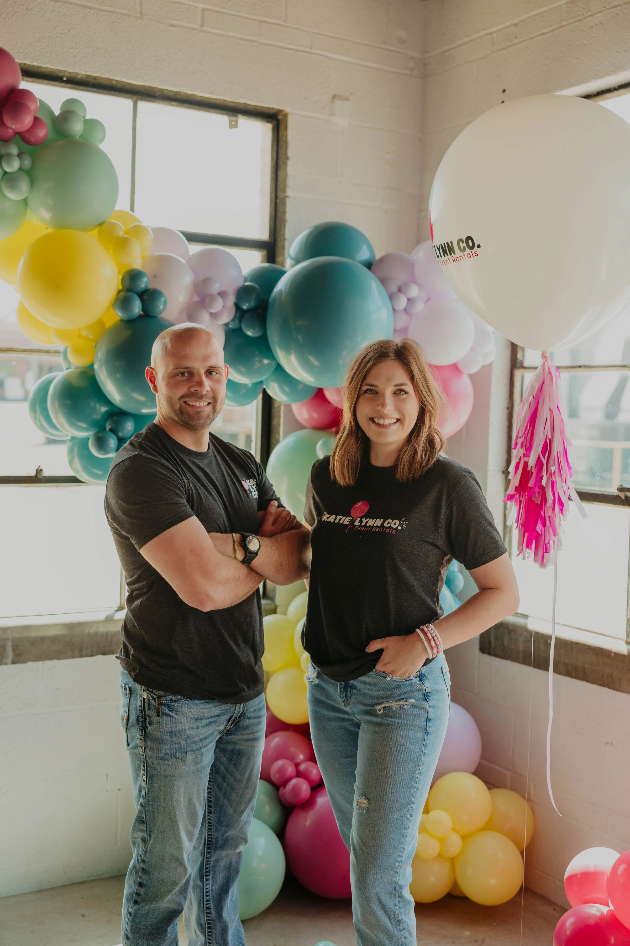 A man and woman standing in front of colorful balloons in a bright indoor setting, smiling at the camera.