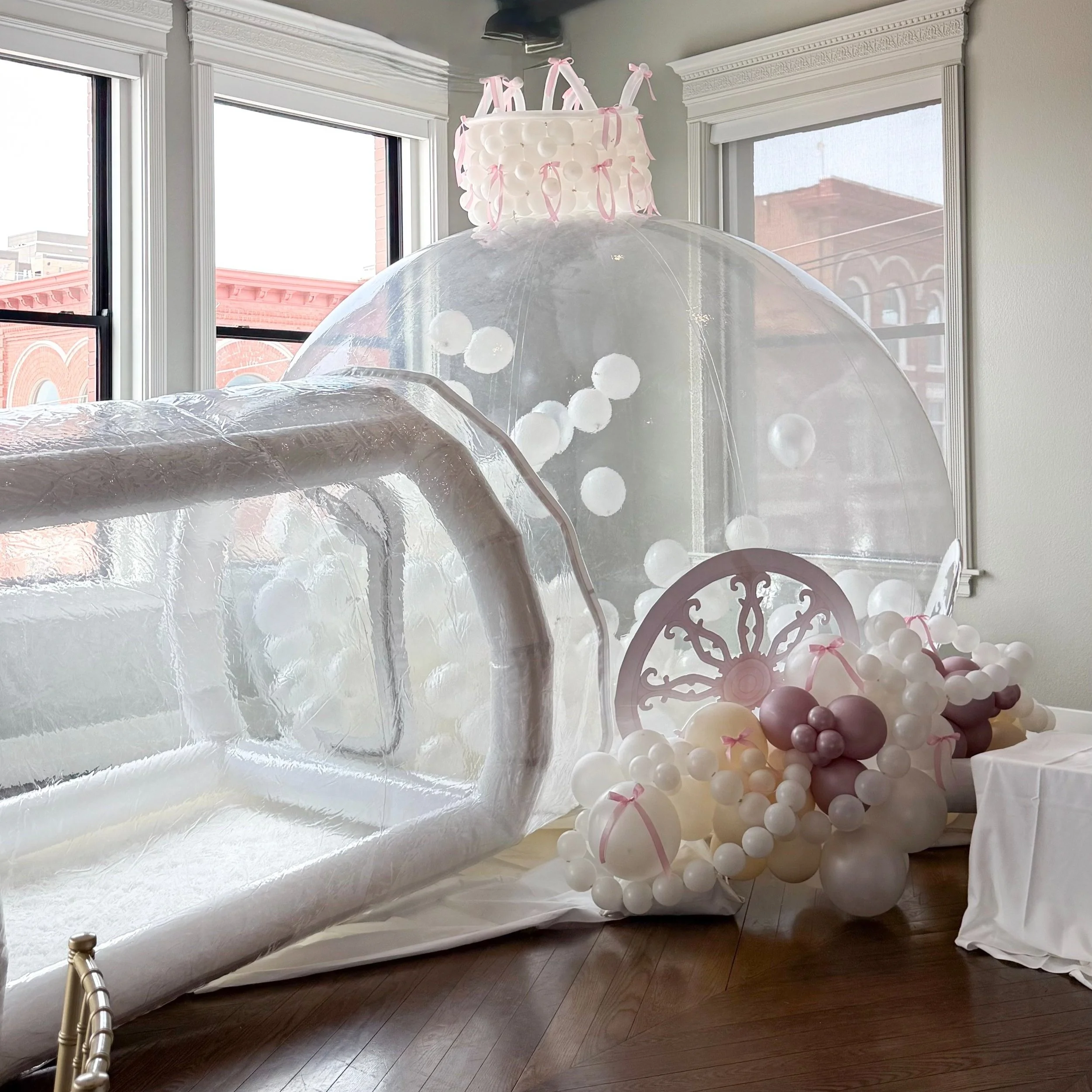 Decorative balloon setup with a transparent bubble tent, balloon arch, and pink ribbon accents, likely intended for a celebration or event. The scene is indoors near windows with a city view.