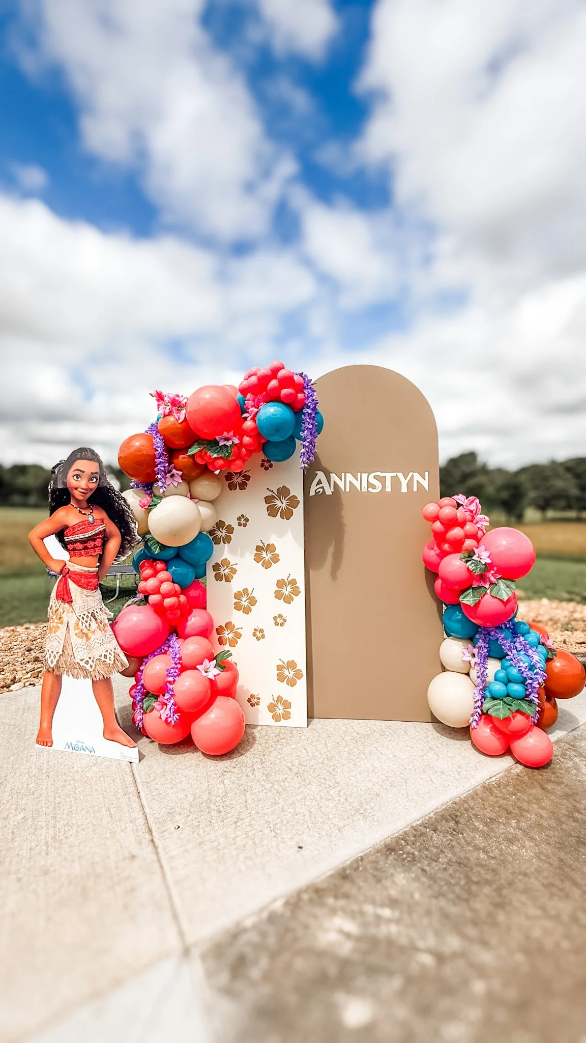 Colorful display with tropical flower and fruit themed balloons around a large sign that says 'Annstyn.' A life-sized cutout of Moana character from Disney Moana stands nearby, set outdoors under a partly cloudy sky.