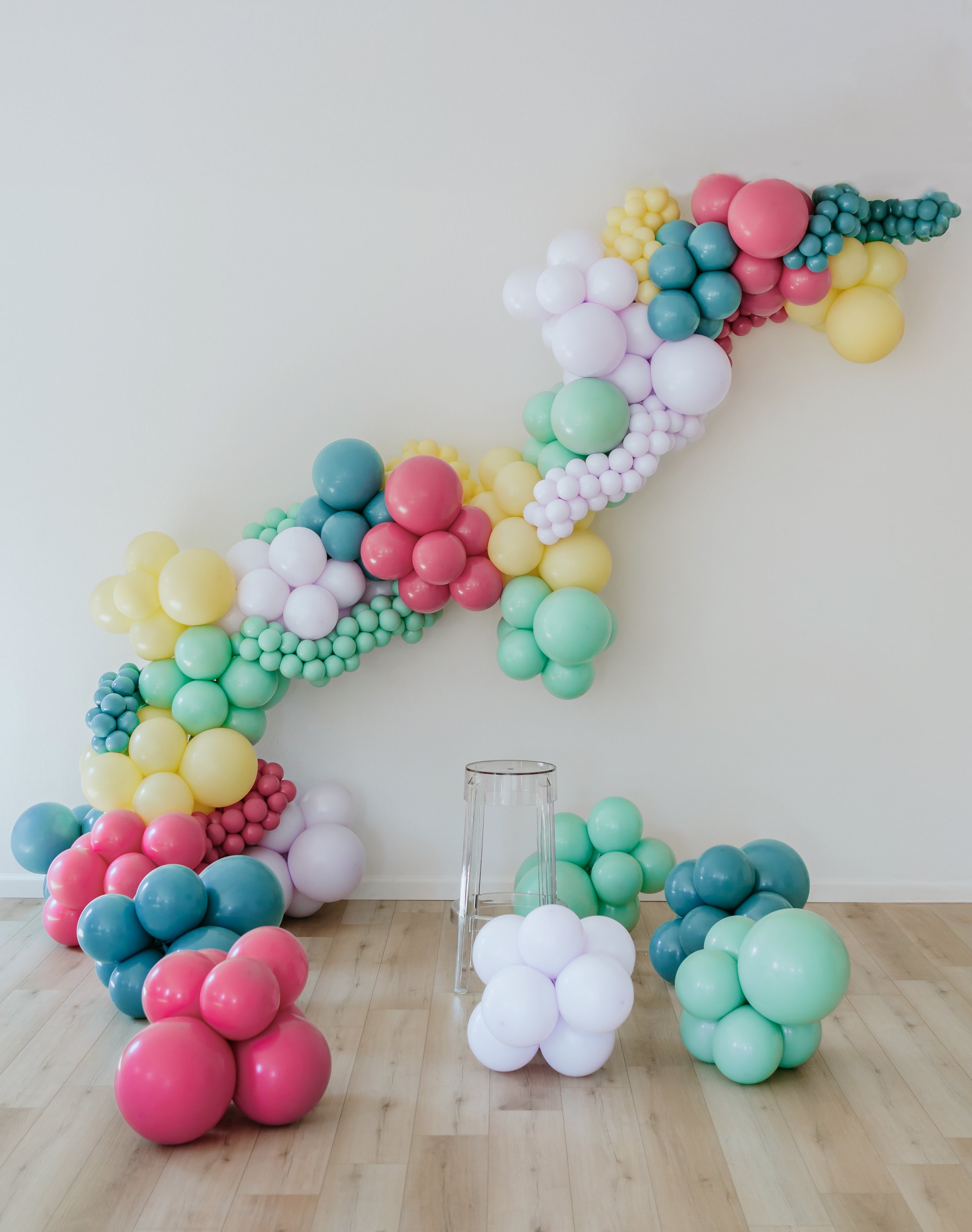 A colorful balloon art installation featuring pink, white, yellow, mint green, and teal balloons in various sizes, arranged against a plain white wall with a wooden floor underneath.