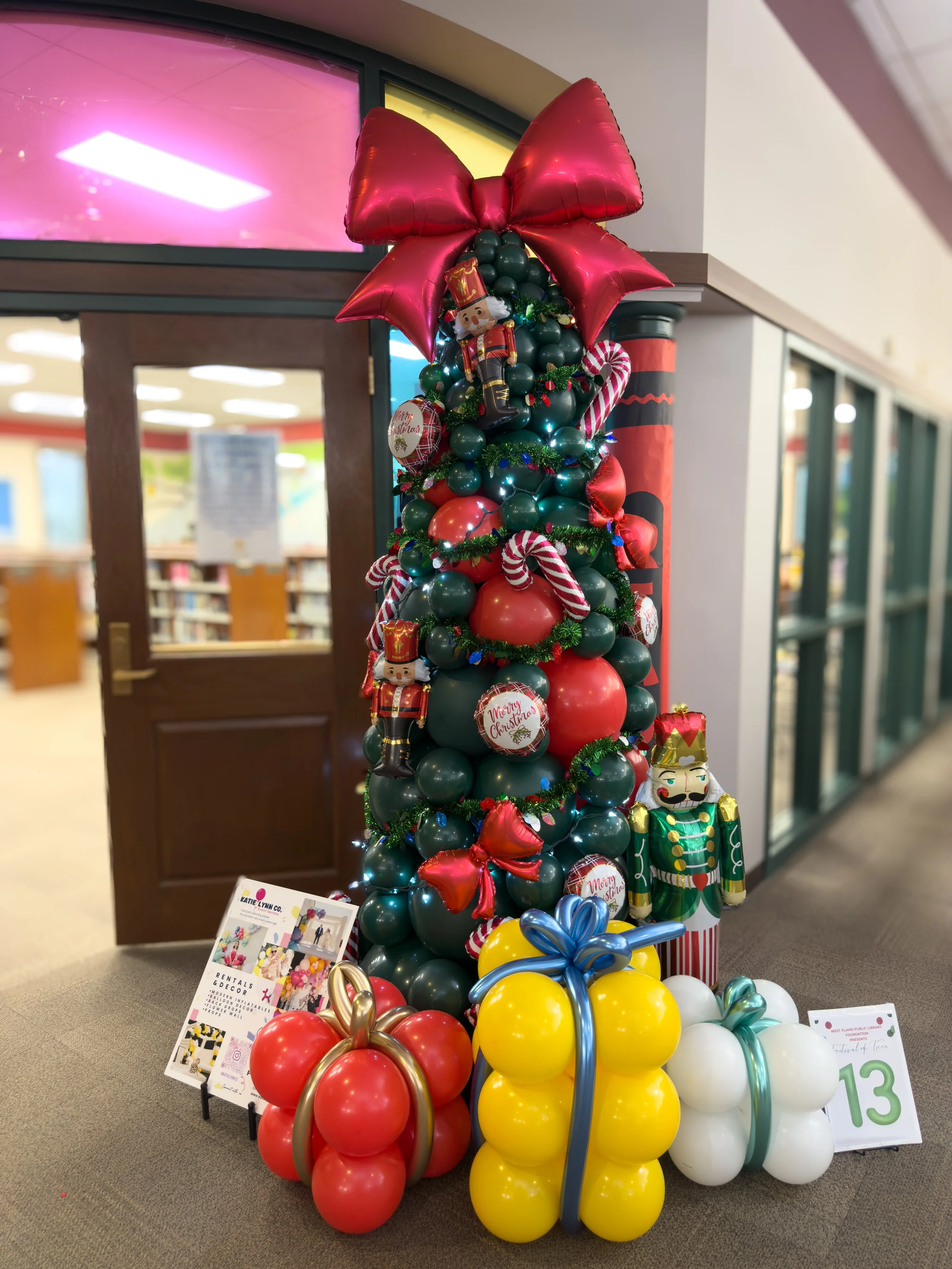 Decorated Christmas tree made of balloons in red, green, yellow, and white, topped with a large red bow. The tree features ornaments including nutcracker figures, candy canes, and round baubles, with some signs and displays nearby.