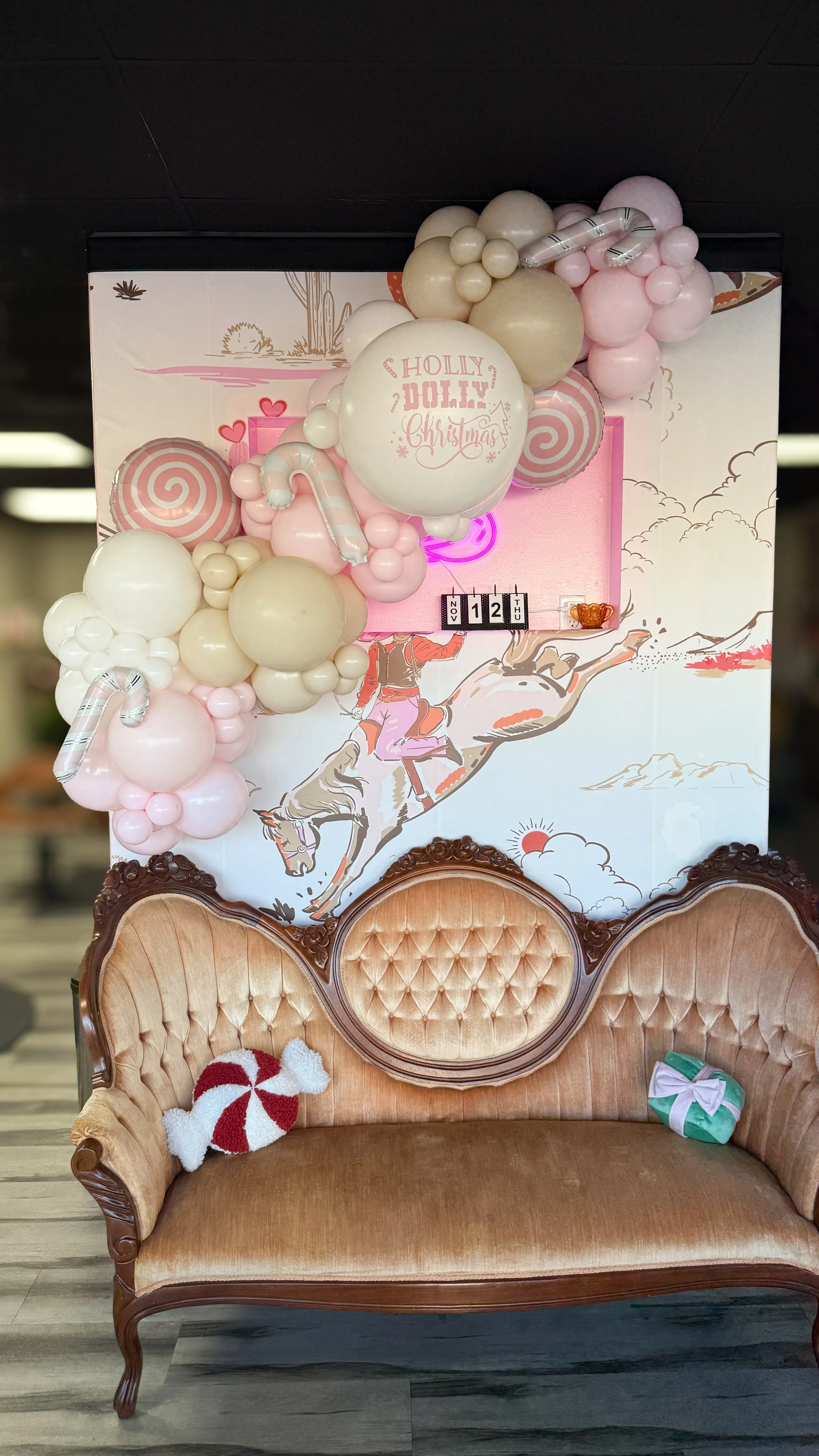 A vintage velvet sofa with a teddy bear holding a peppermint cushion and a wrapped gift on the armrest, in front of a decorated wall with balloons and a holiday-themed illustration.