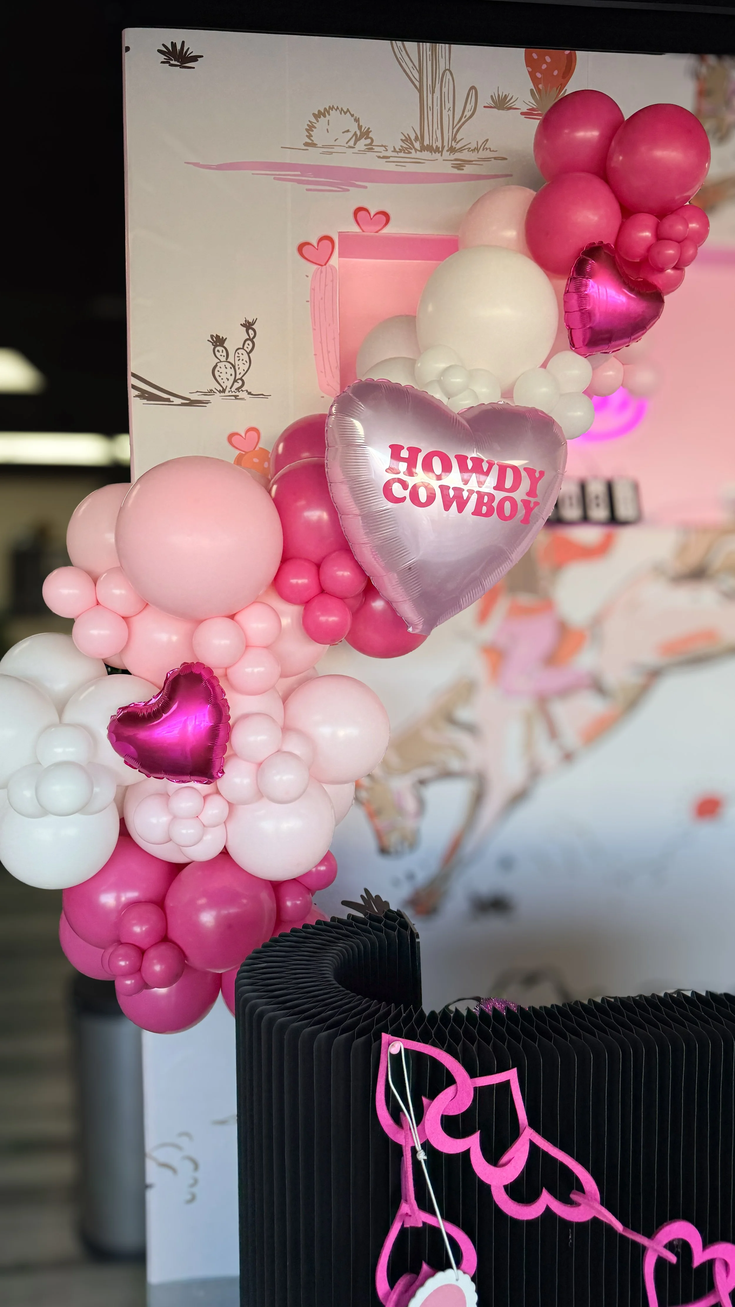Pink, white, and purple balloons forming a decorative arch with heart-shaped balloons and a silver balloon that says 'Howdy Cowboy' in pink letters. The background has a cowboy-themed mural with cacti and desert scenery.