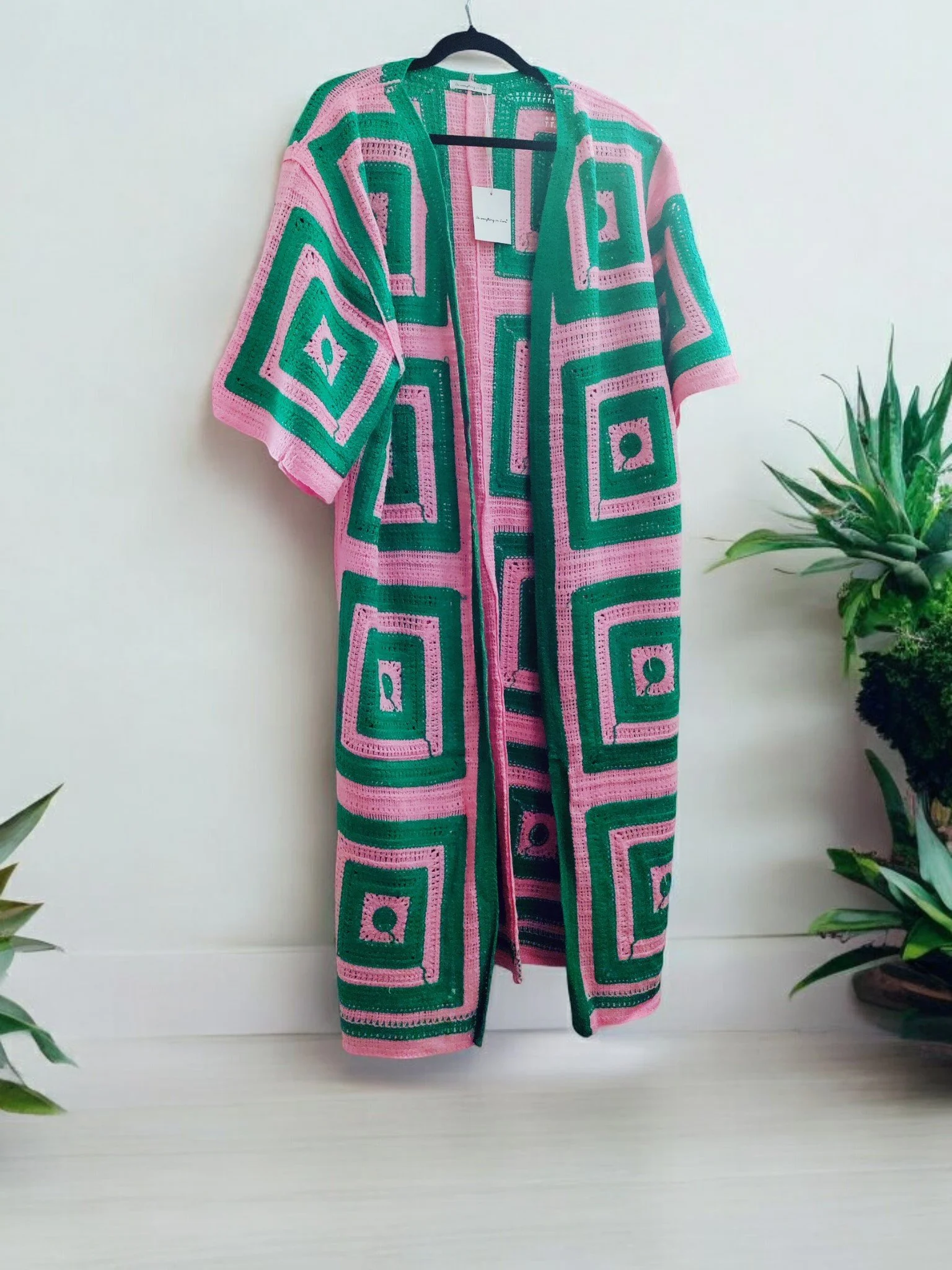 Crocheted kimono with pink and green geometric patterns hanging on a black hanger against a white wall, surrounded by green plants.