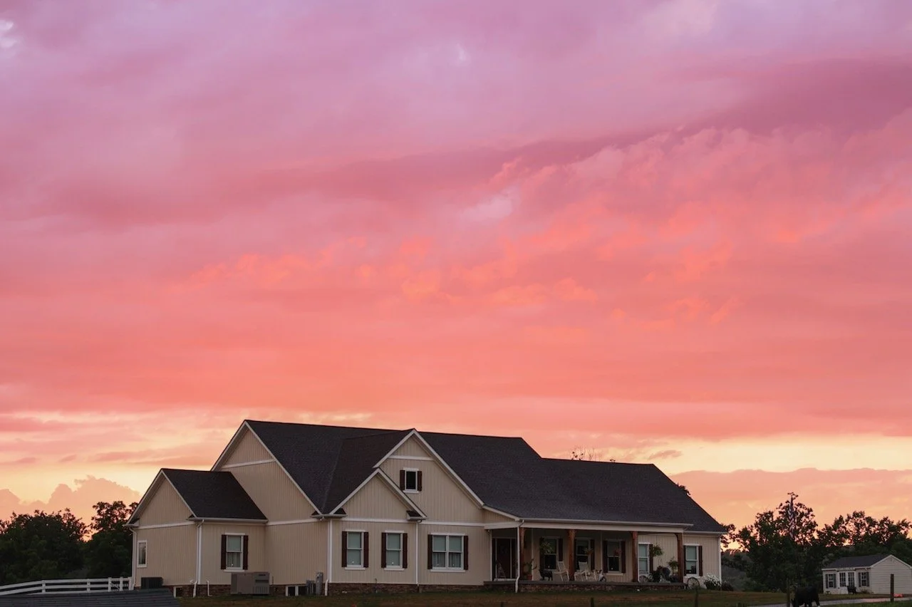 A large house with a porch under a colorful pink and purple sunset sky, with trees and a smaller house in the background.