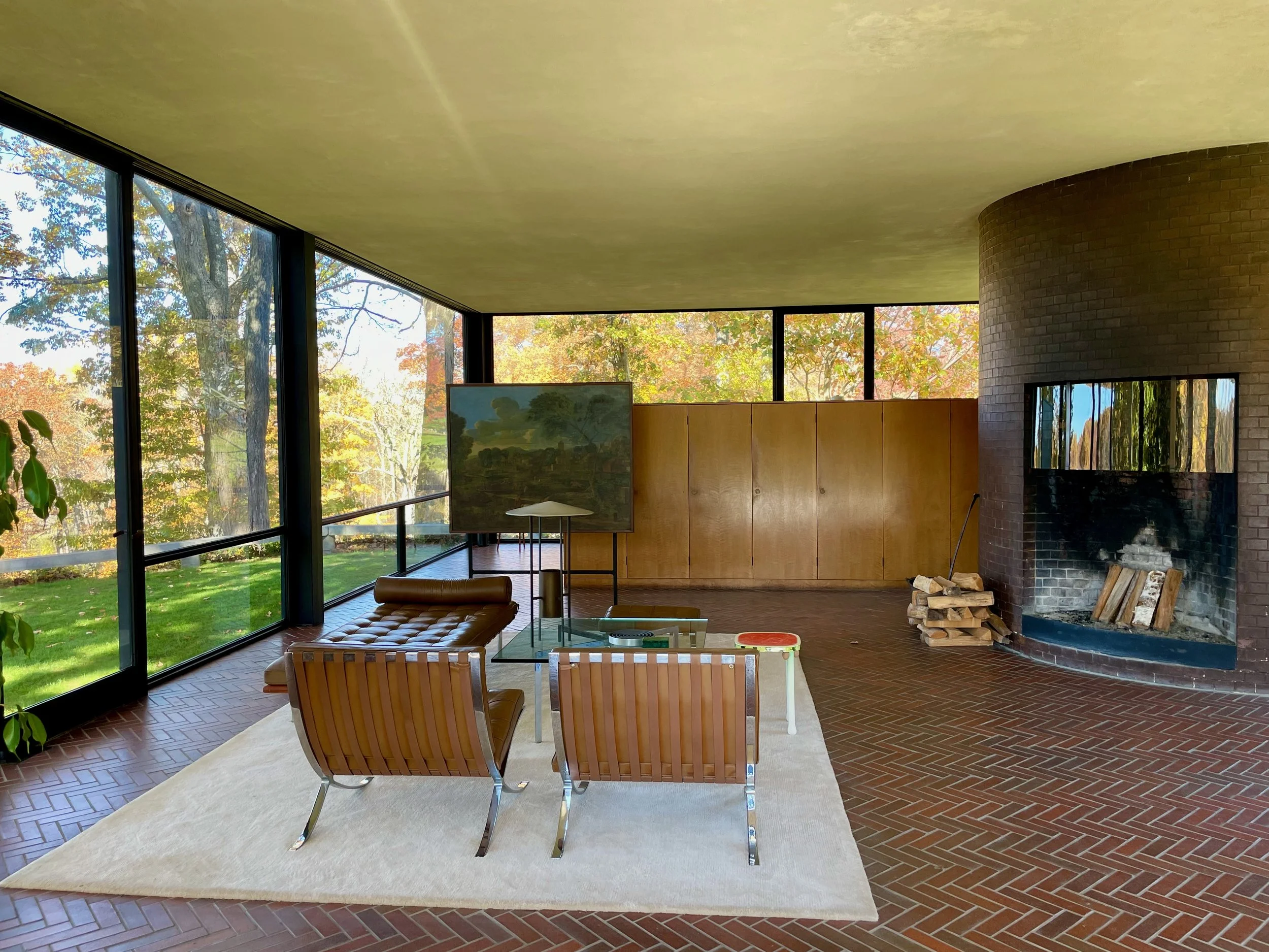 A view of Philip Johnson's Glass House showing autumn trees outside, a fireplace with stacked firewood, a beige rug, two wooden chairs, and a small table. There's a painting on a stand and a beige leather lounge chair.