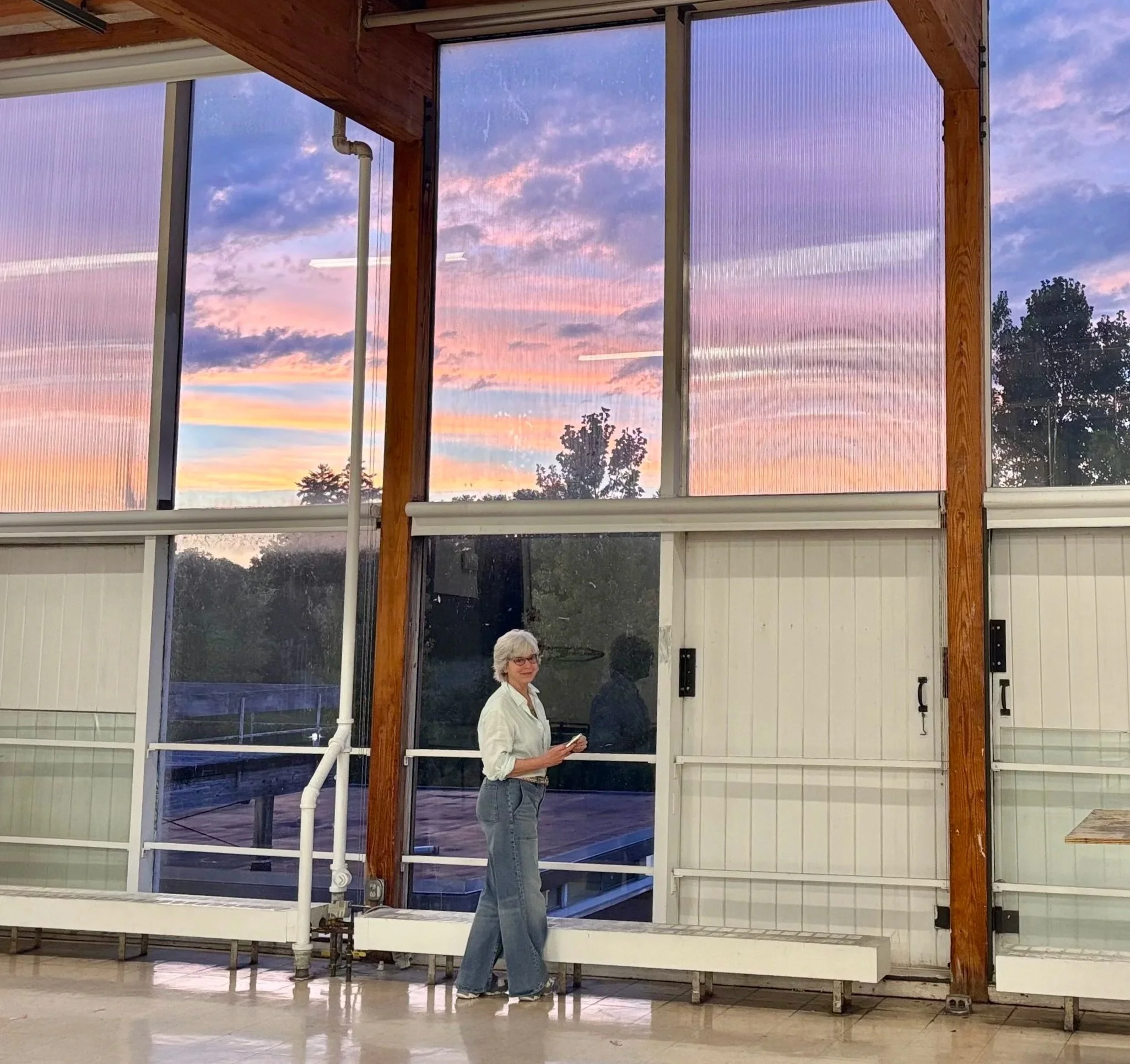 A woman with silvery hair and glasses, wearing a pale blue green shirt and blue jeans, standing indoors near large glass windows during sunset, with a view of trees outside.