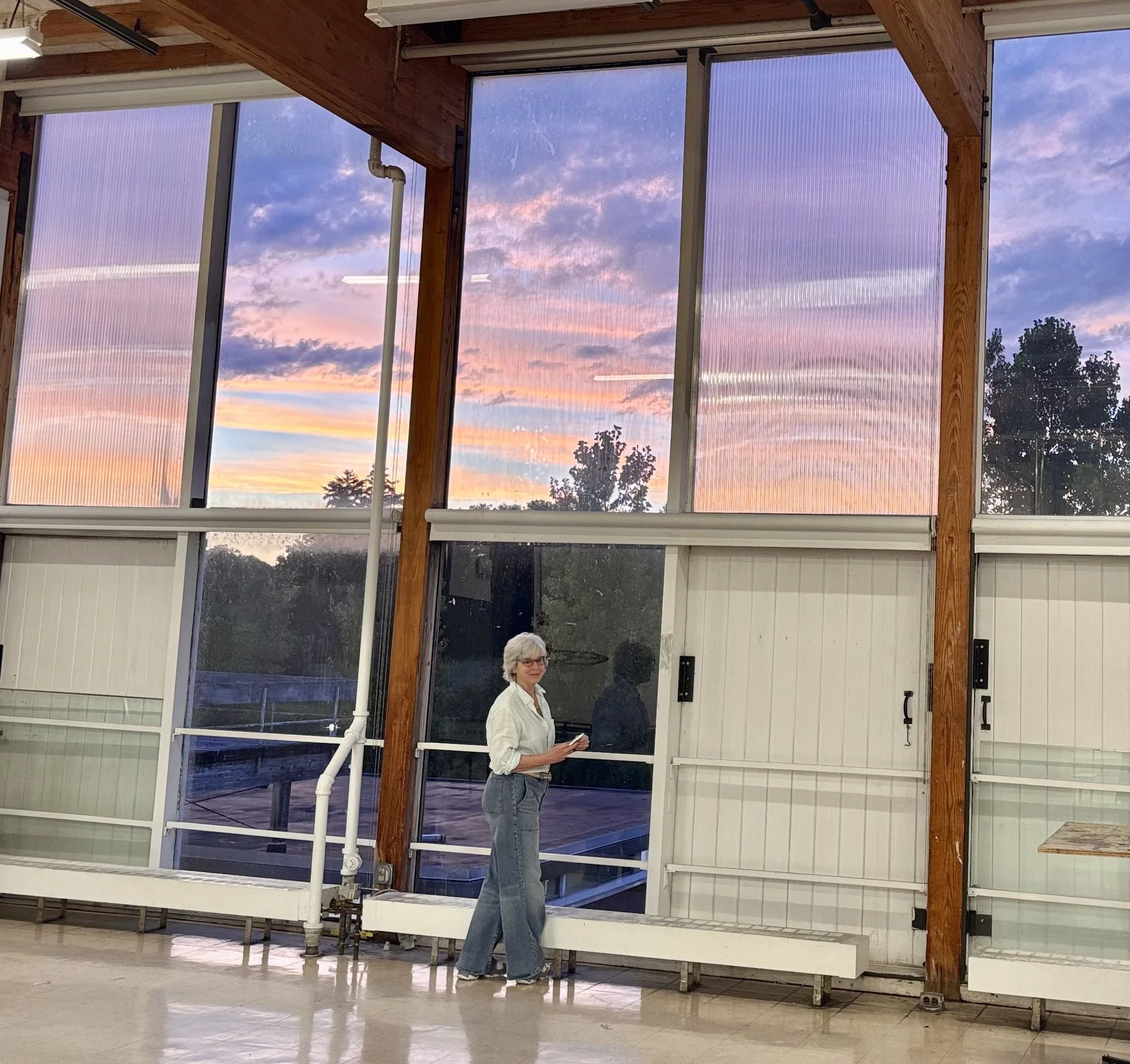 An indoor space with large windows showing a colorful sunset sky, with a woman wearing glasses, a white shirt, and jeans standing inside near the windows.