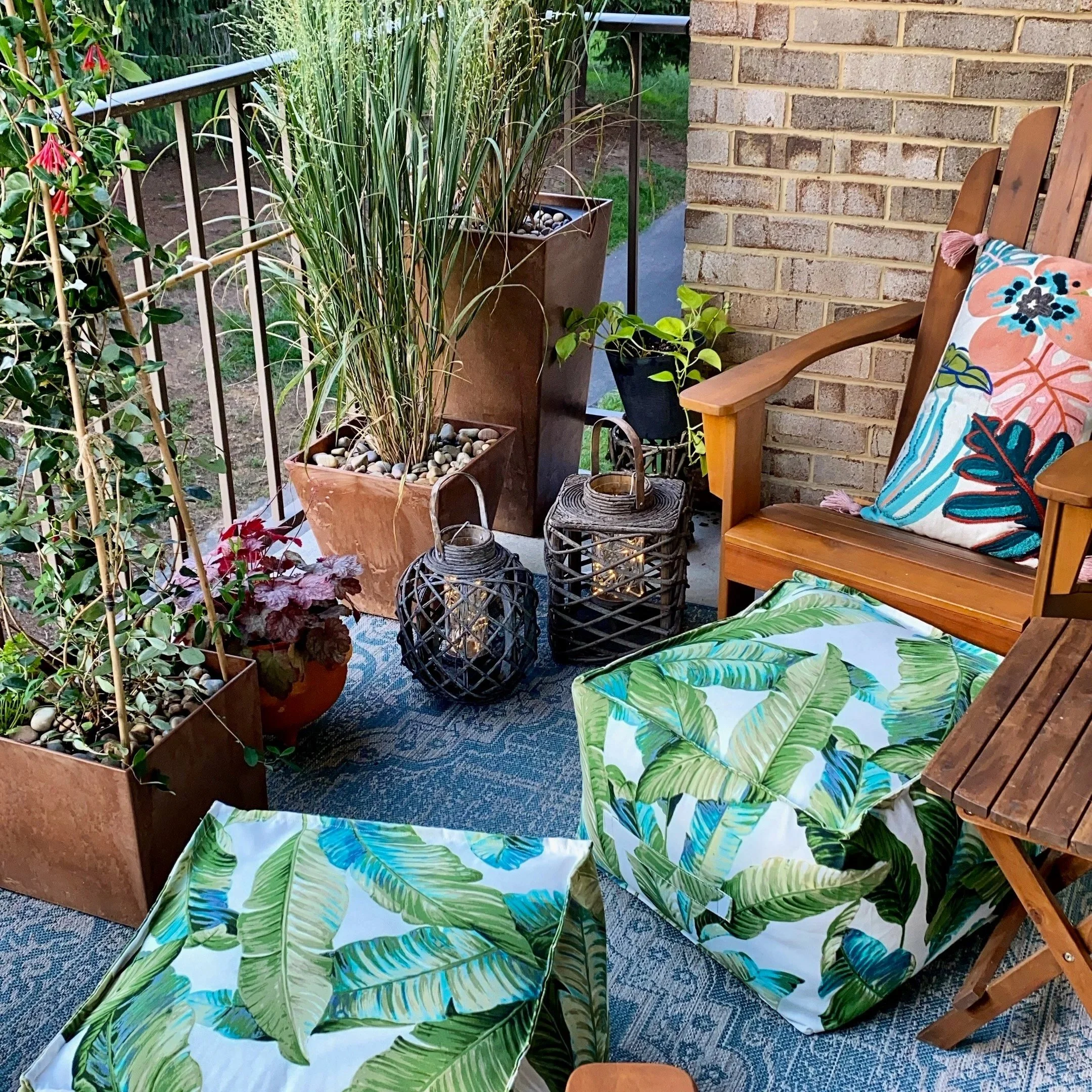 A cozy balcony with potted plants, wooden furniture, a colorful floral throw pillow, and tropical leaf-patterned fabric ottomans. The balcony has a brick wall and is decorated with lanterns, a rug, and side table.