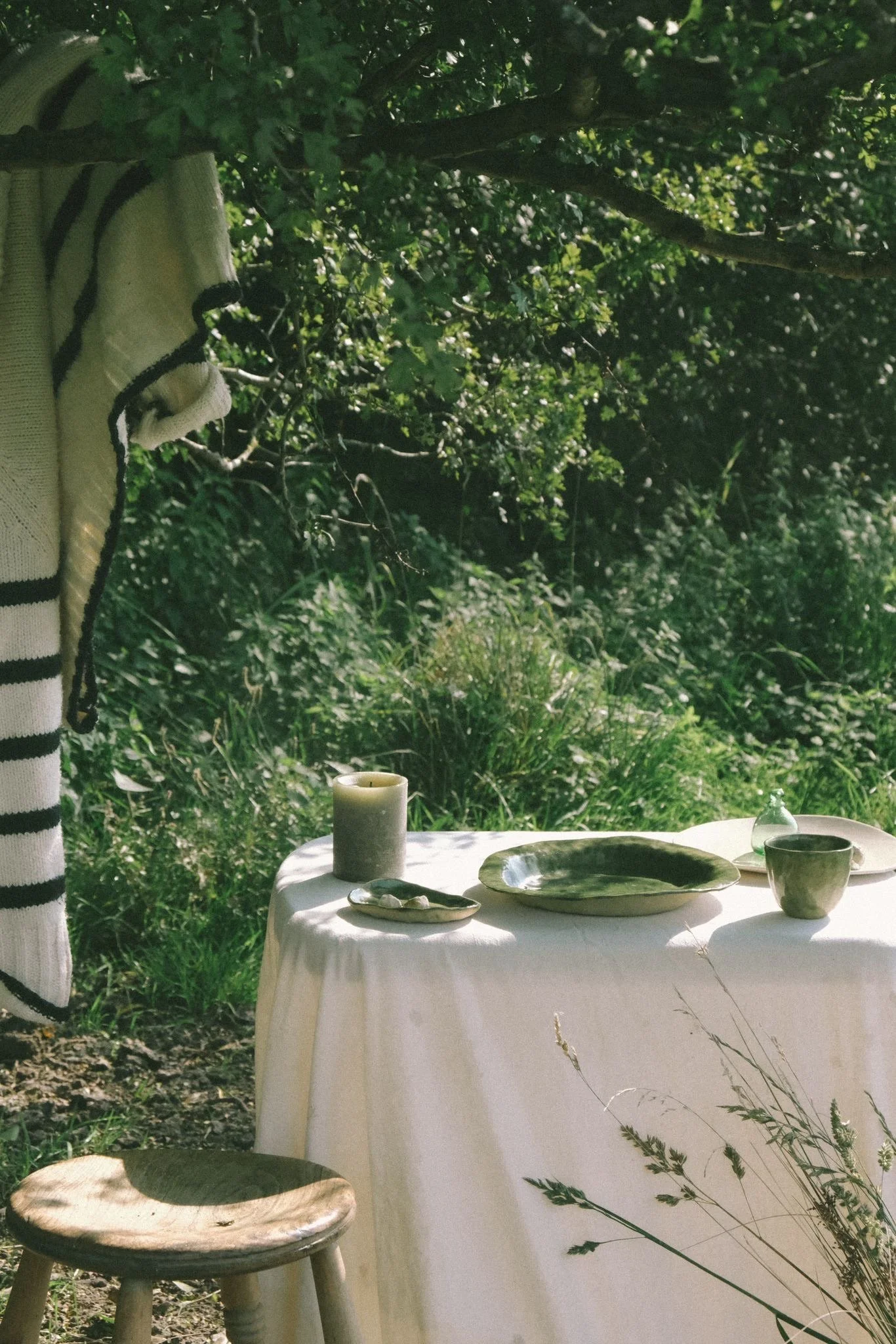 An outdoor table with a white tablecloth set with green dishware, a candle, and a small decorative bottle, surrounded by greenery.