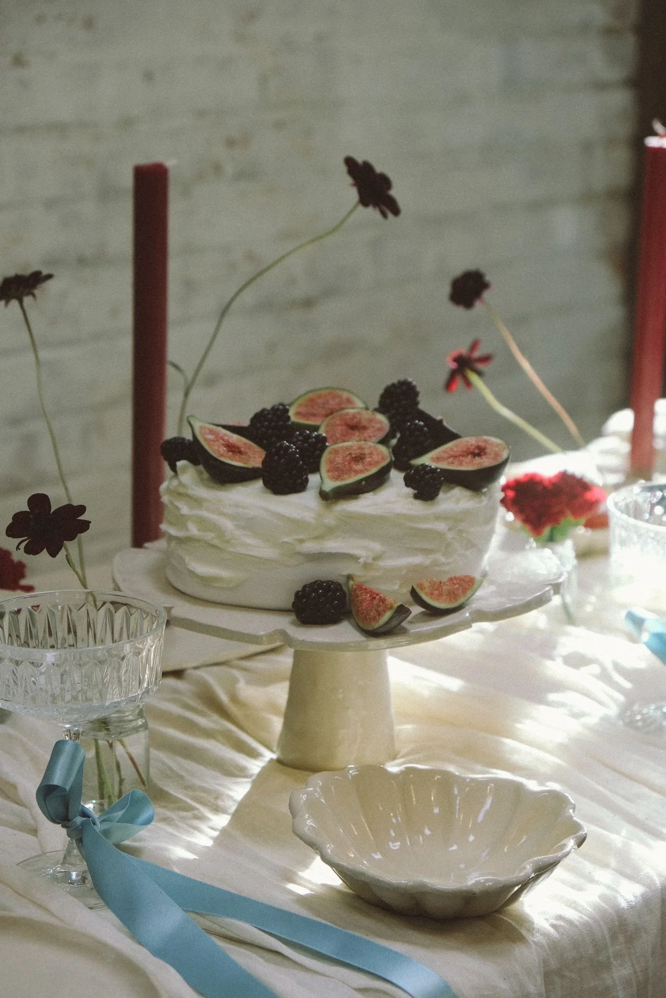 A white frosted cake decorated with fresh figs and blackberries on a cake stand, surrounded by other glassware and a light-colored tablecloth.