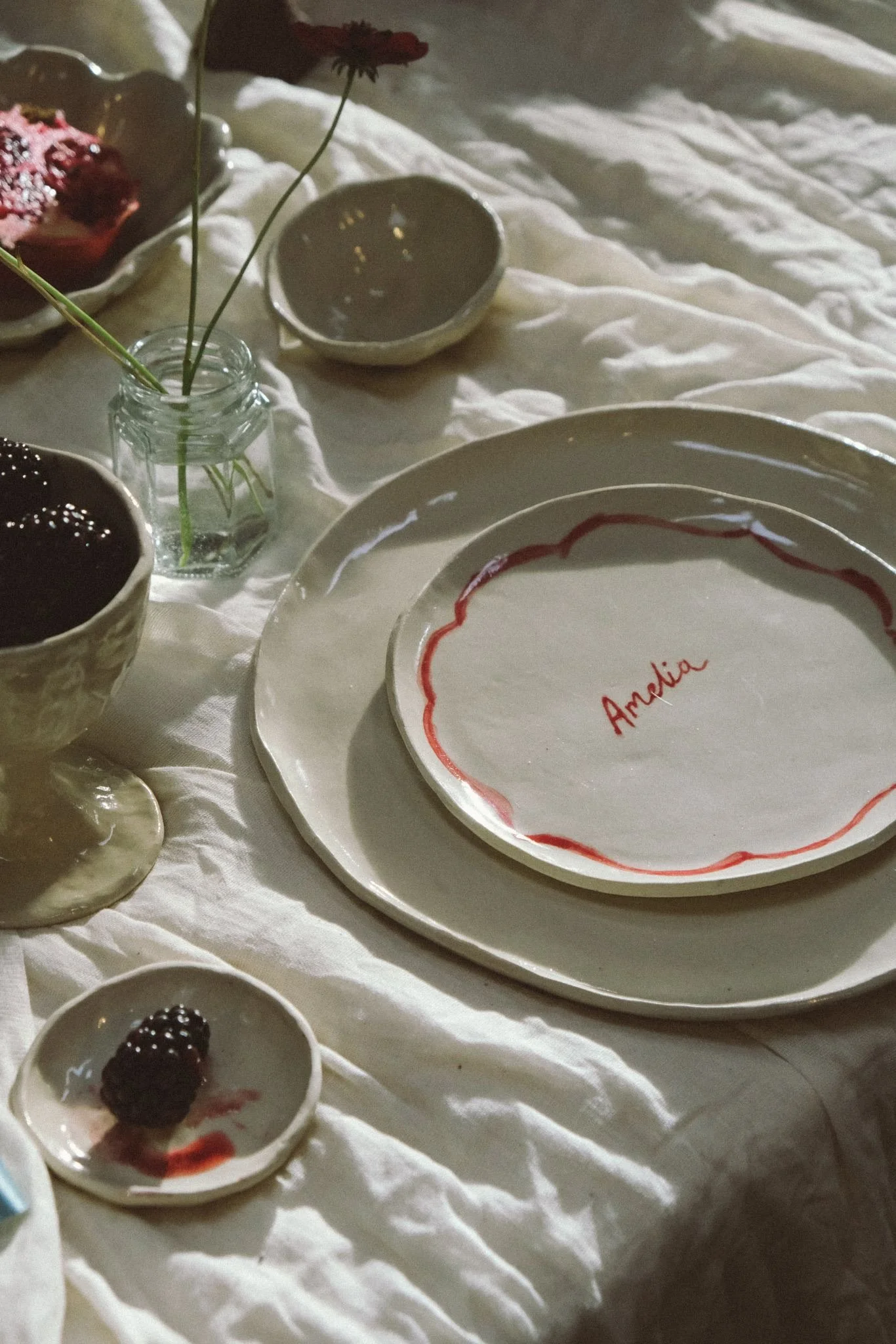 A white tablecloth with handmade ceramic plates, a small vase with a single red flower, and various small bowls and dishes, some with berries and drizzles of sauce, in a rustic, cozy setting.