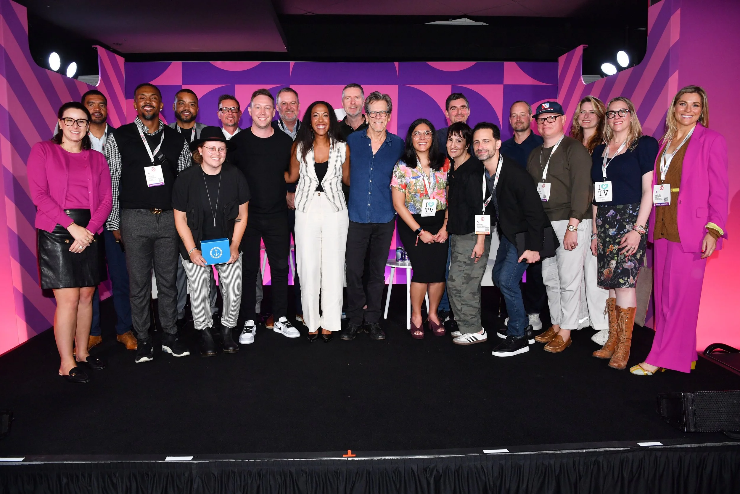 A diverse group of smiling adults stand on a stage in front of a bright pink and purple background