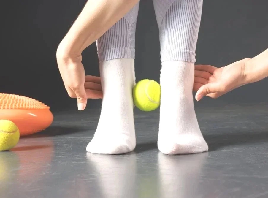 “Adult feet guided through proprioceptive training, using a ball between the ankles to improve stability and load transfer.”