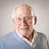 Elderly man with glasses smiling against gray background.