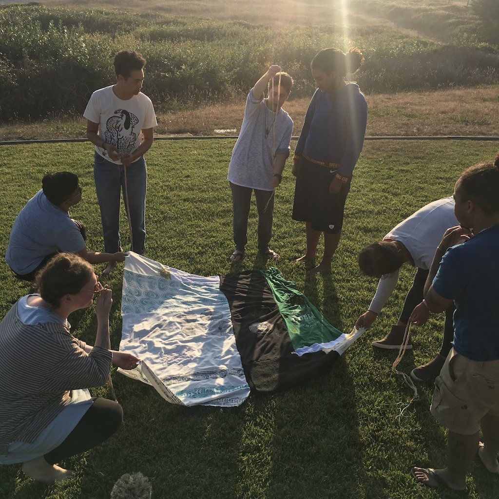A group of of queer young people with brown, tawny and light skin gather around a multi-colored cloth in field as the sun sets.