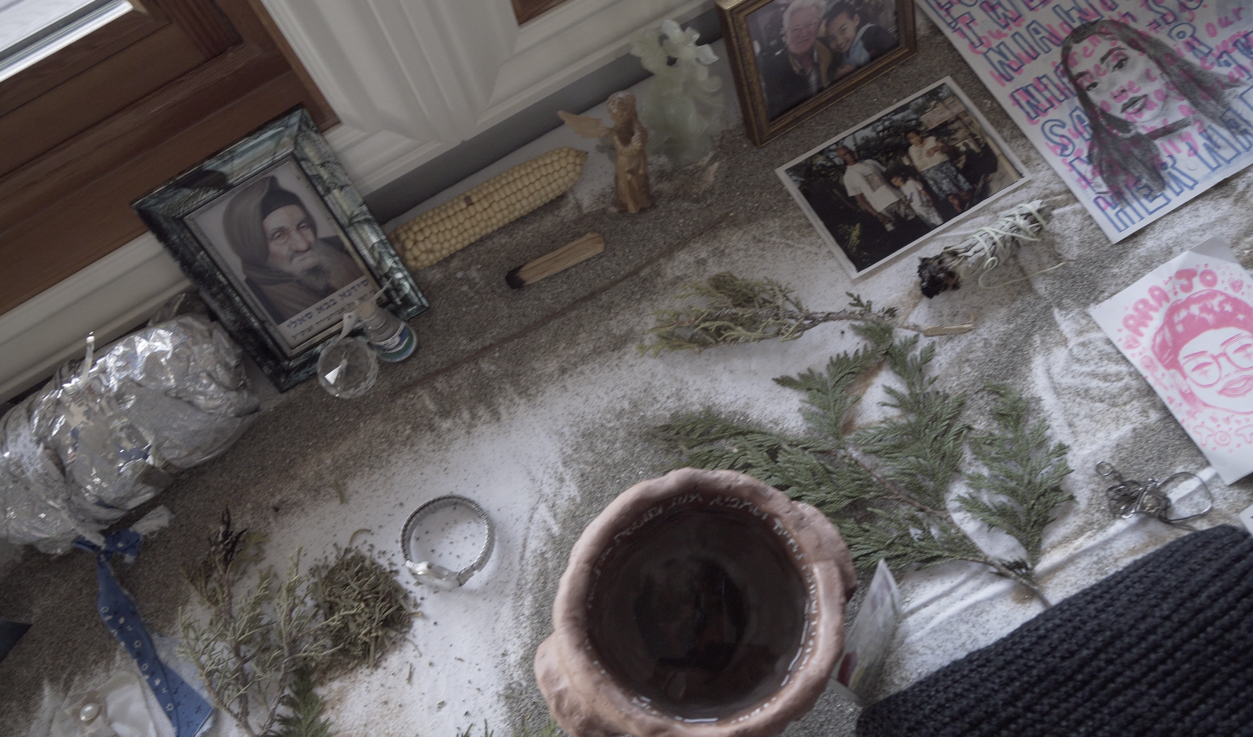 A ceramic pot holding water rests at the center of an artfully arranged altar. The altar also holds pictures of ancestors and plant clippings.