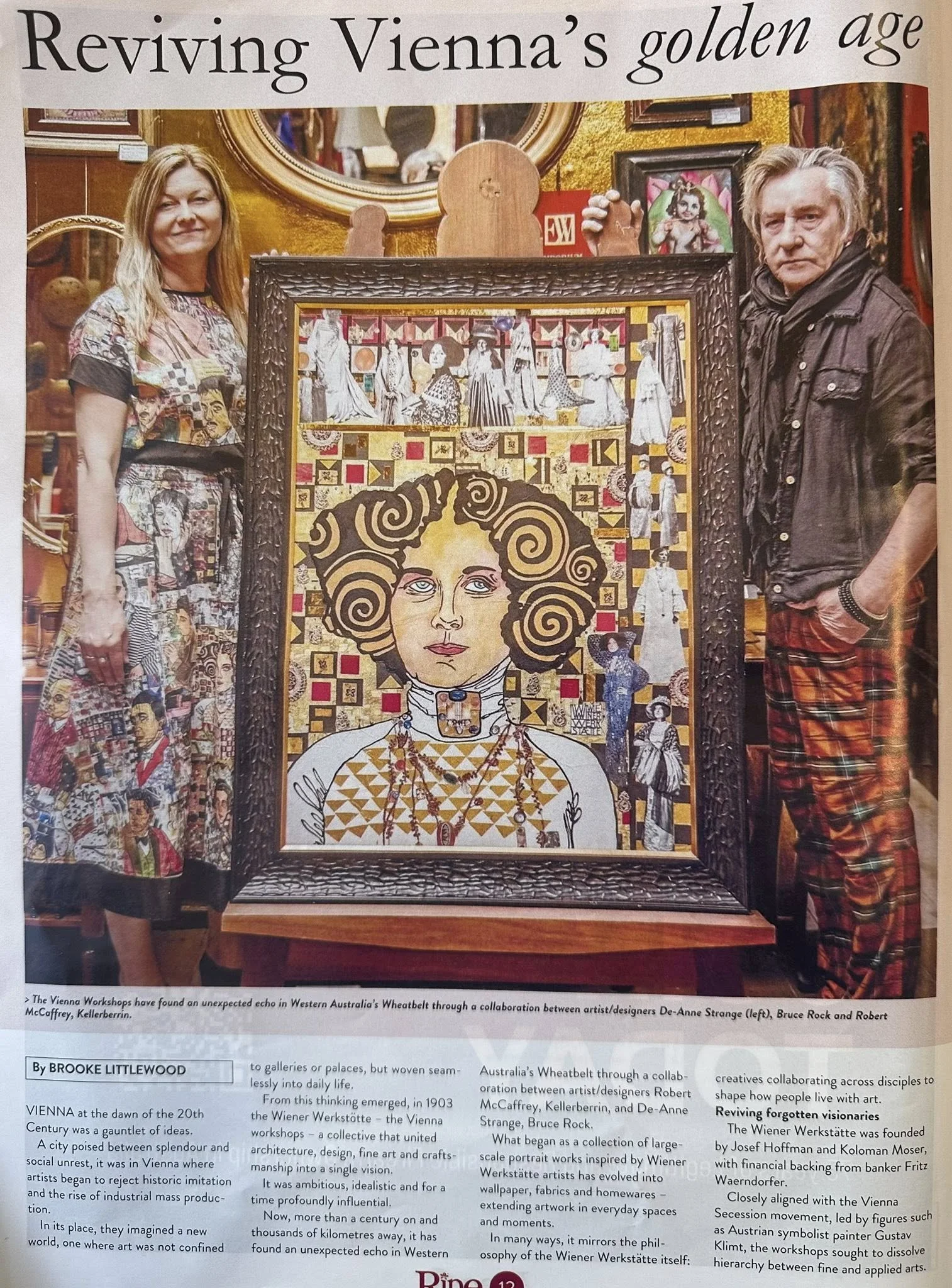 A photo of two people standing behind an artwork piece in a gallery. The woman on the left has blonde hair, is smiling, and is wearing a patterned dress. The man on the right has blonde or gray hair, a serious expression, and is wearing a black jacket with plaid pants. The artwork features a collage with a woman's face, swirling hair, and various patterns. The background shows gold and colorful framed art pieces.