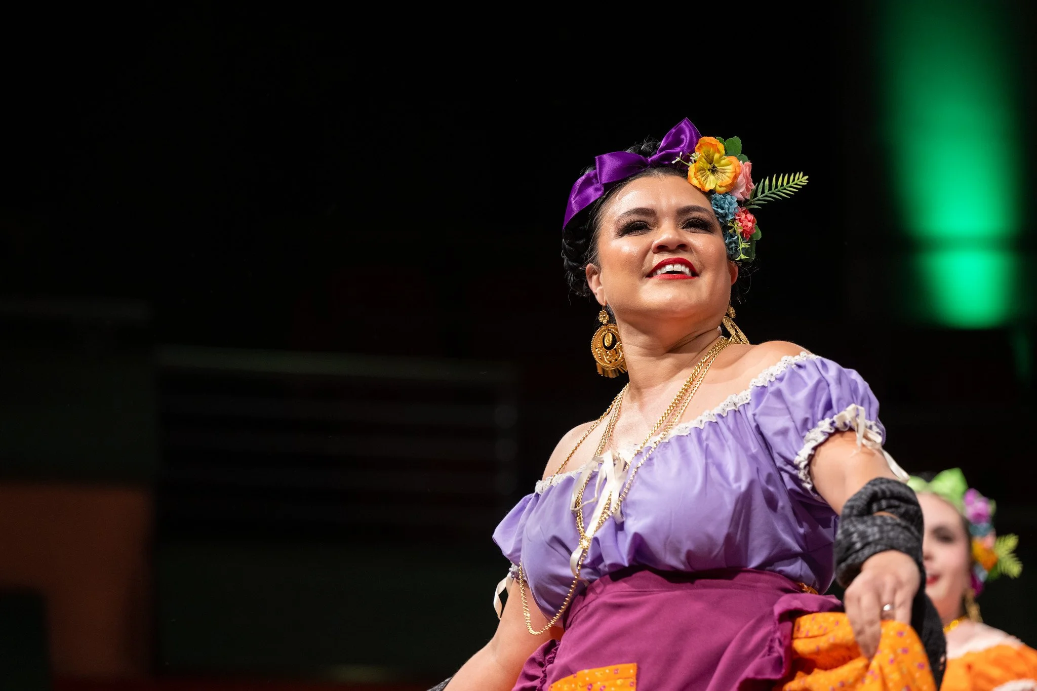 Woman dressed in colorful traditional Mexican attire, wearing purple and orange clothing and floral headpiece, performing a dance.