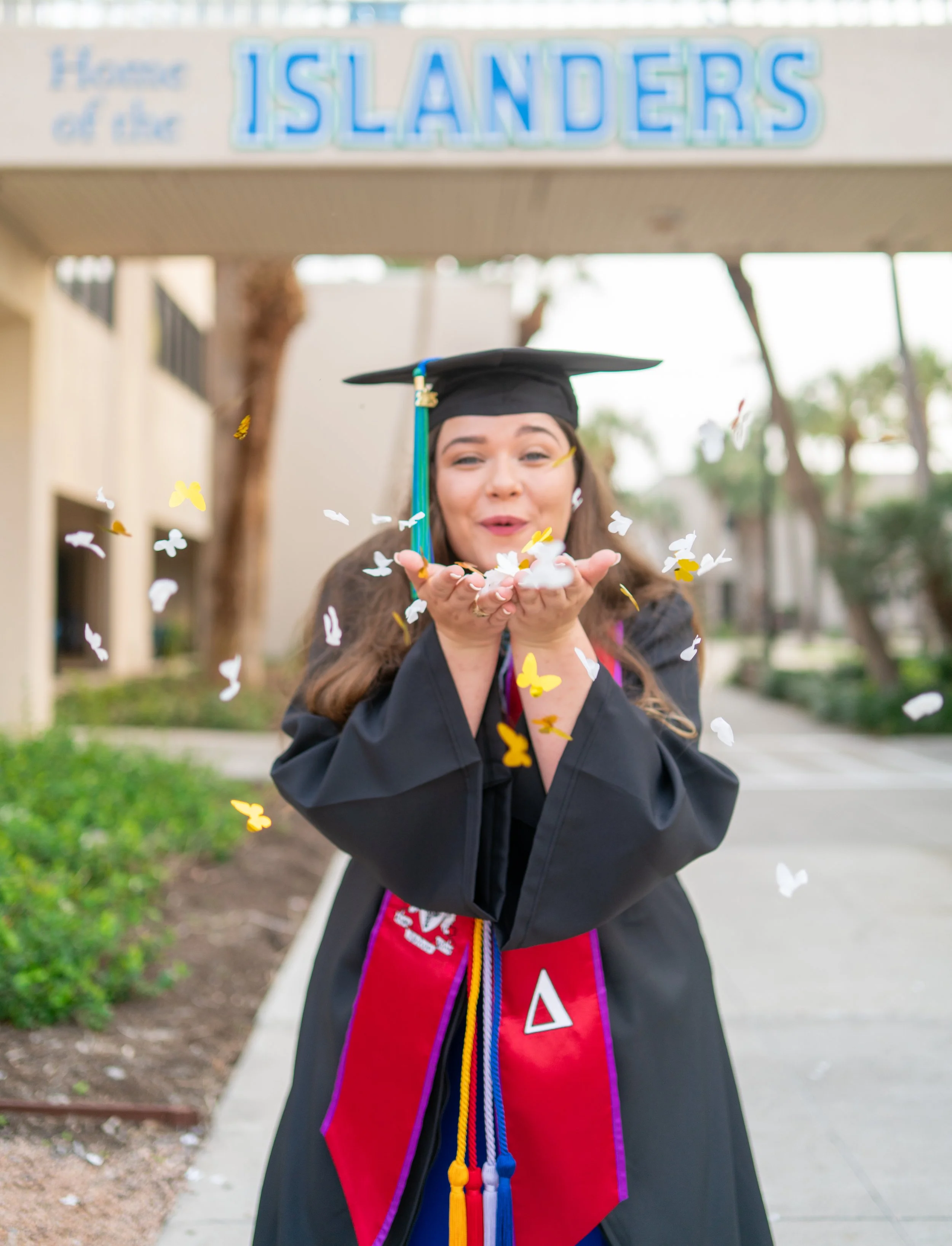 Woman in graduation gown and cap blowing confetti outdoors near a sign that reads "Islanders".