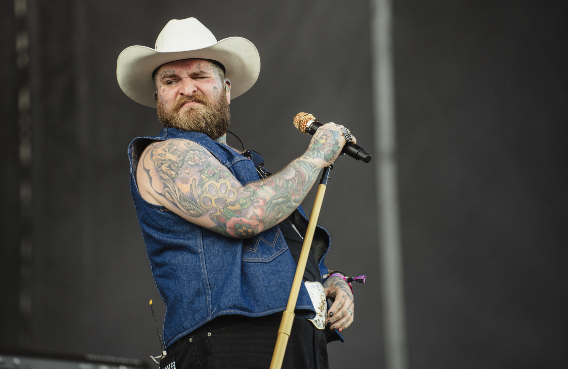 A bearded man wearing a cowboy hat and sleeveless denim shirt, holding a microphone, stands on stage with tattoos on his arms, and appears to be performing or speaking.