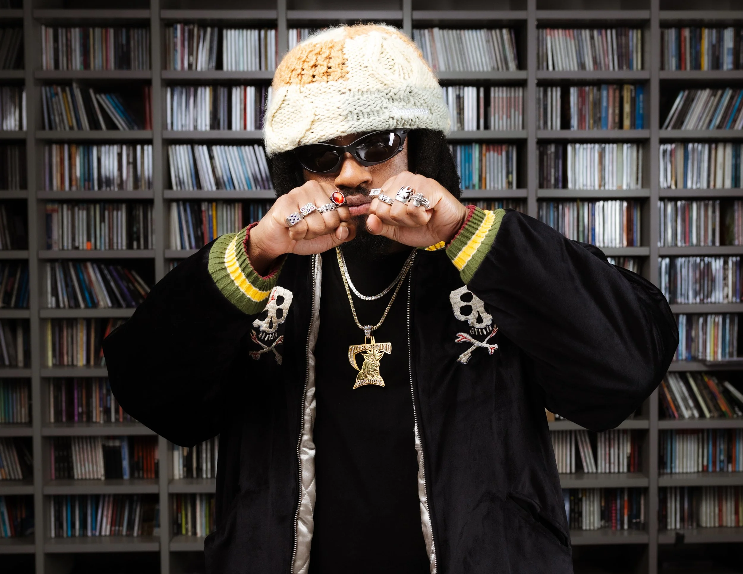 A man wearing a colorful knit beanie, sunglasses, a black jacket with skull and crossbones embroidery, and multiple rings and necklaces, standing in front of a wall filled with CDs.