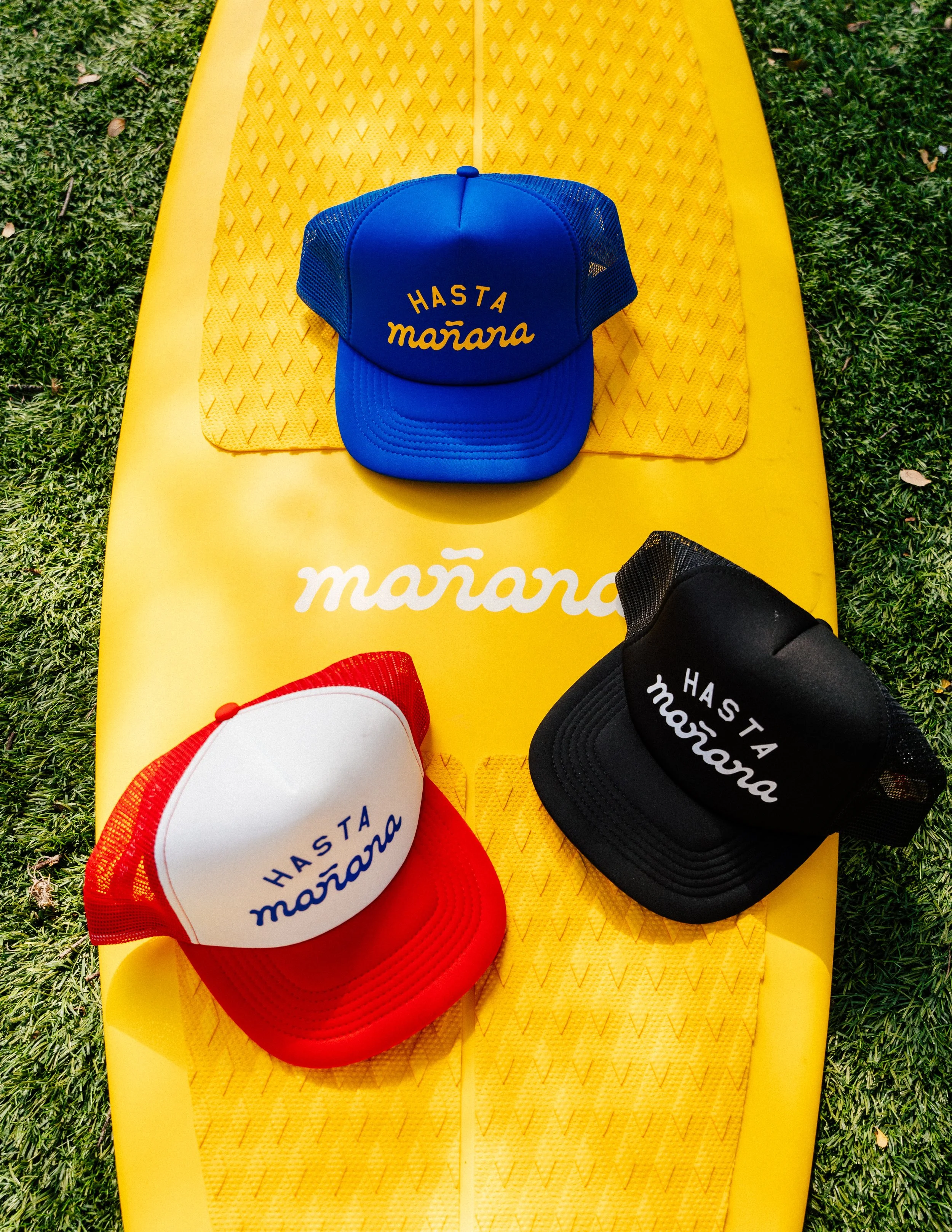 Three trucker hats with the phrase 'Hasta Mañana' in different colors: blue, red and white, black, displayed on a yellow paddleboard outdoors on grass.